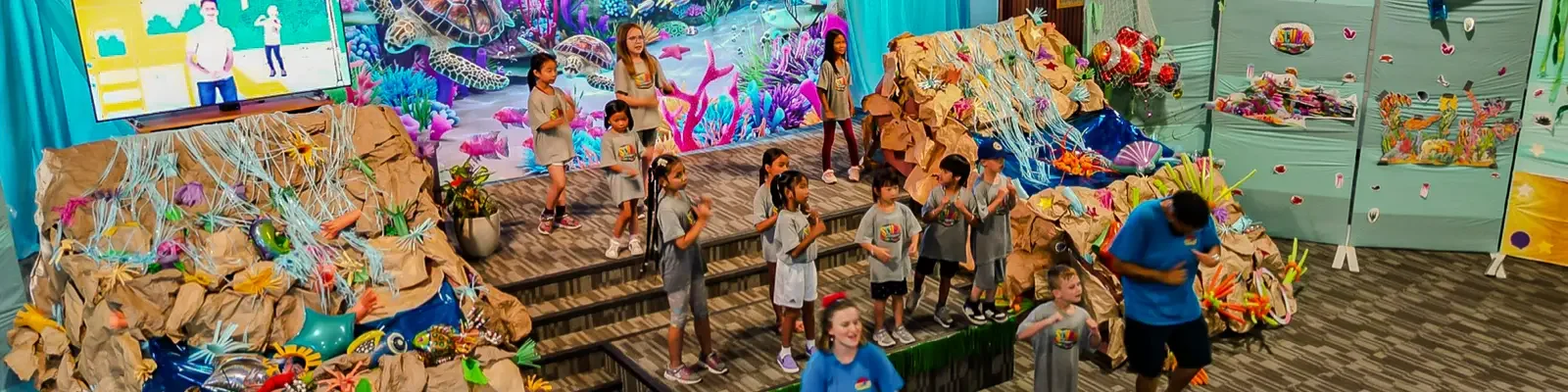 Children performing on a stage with an ocean-themed backdrop, surrounded by paper-mâché mountains and sea life decorations