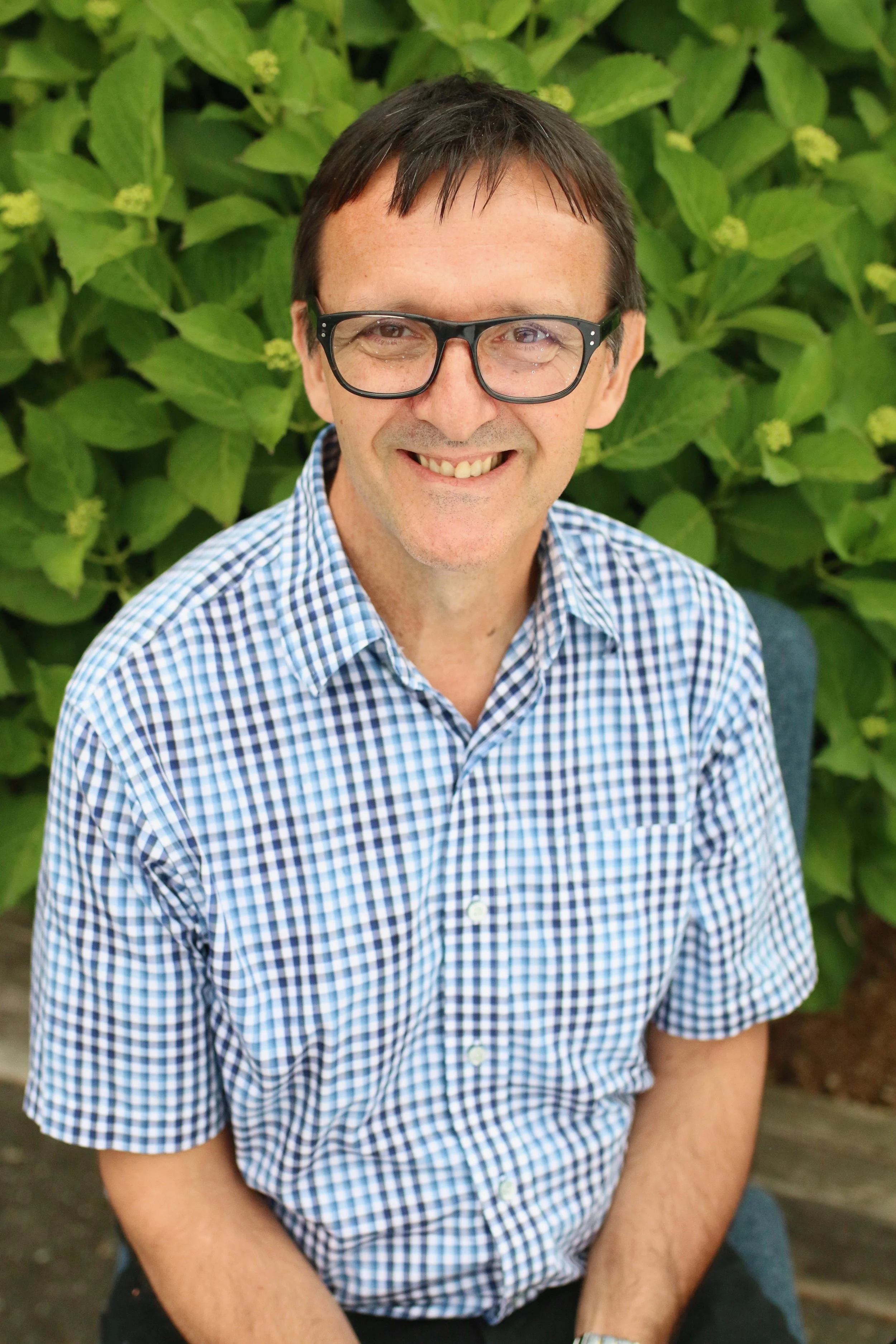 Smiling man with short dark hair, wearing glasses and a blue checkered shirt, sitting outdoors in front of green leafy plants.