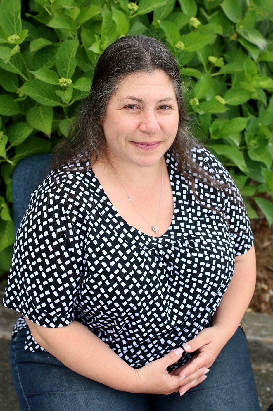 A woman with long brown hair and a slight smile, sitting outdoors in front of green foliage, wearing a black and white patterned top and jeans.