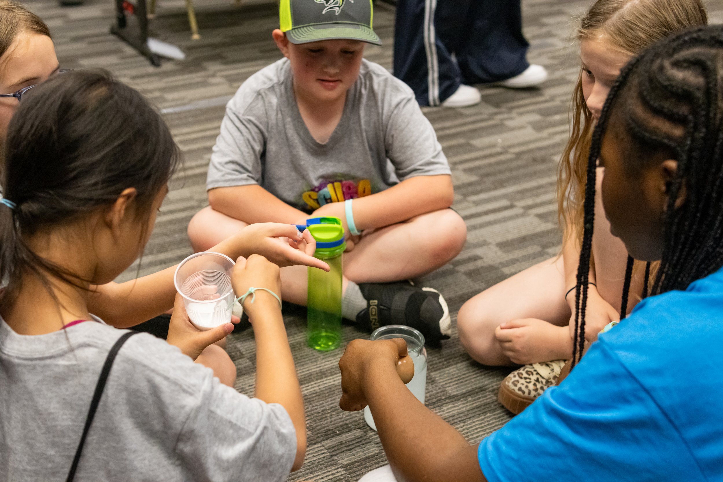 Group of children sitting on a carpeted floor, engaging in a science activity with containers and liquids.