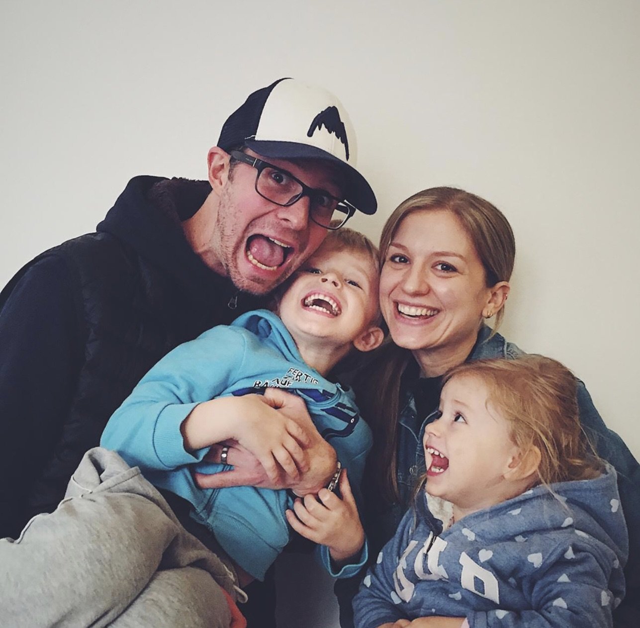 A happy family of five, two adults and three children, smiling and laughing together indoors against a plain wall.