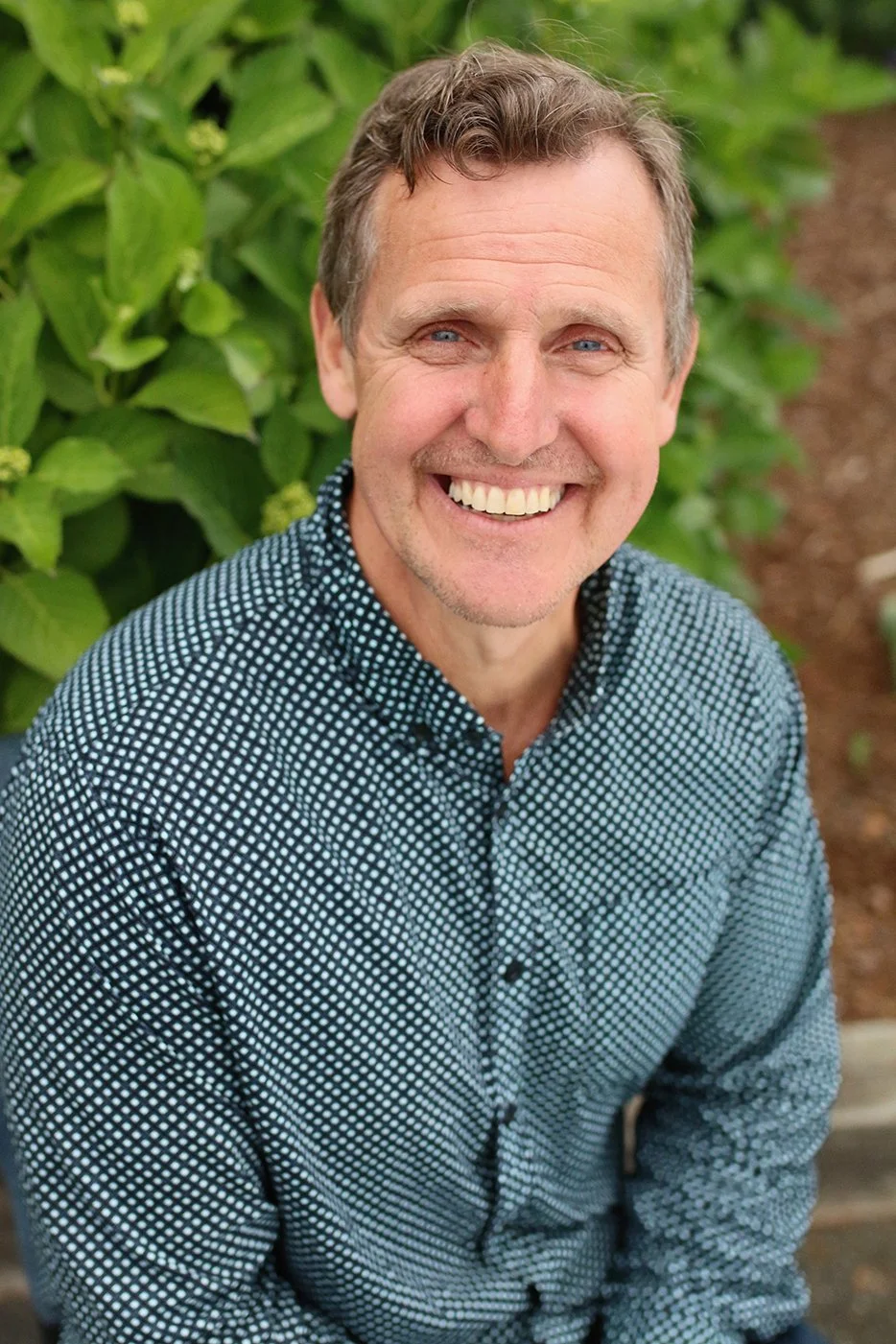 A smiling middle-aged man with short brown hair and blue eyes, wearing a black and white patterned button-up shirt, outdoors with green leafy plants in the background.