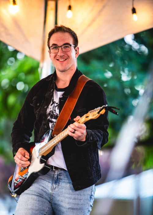 Young man smiling and playing an electric guitar outdoors during evening with string lights overhead.