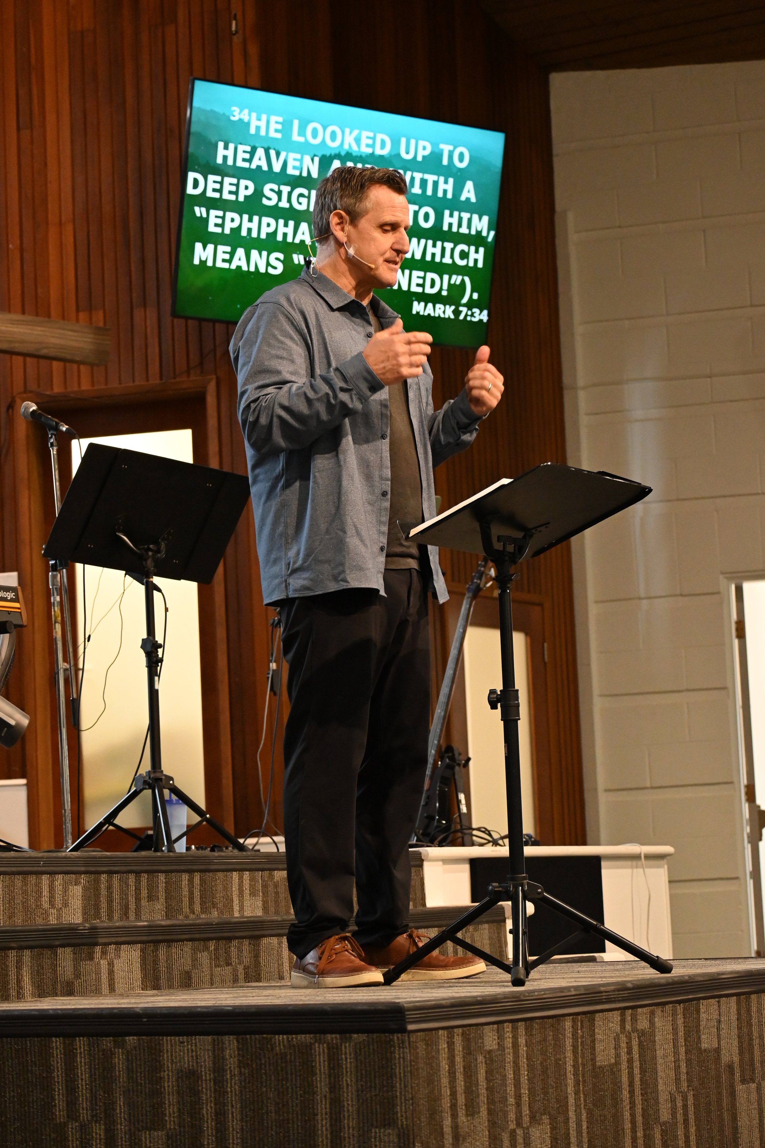 A man giving a speech at a church or auditorium, standing behind a music stand on a wooden stage with a large screen displaying a Bible verse behind him.