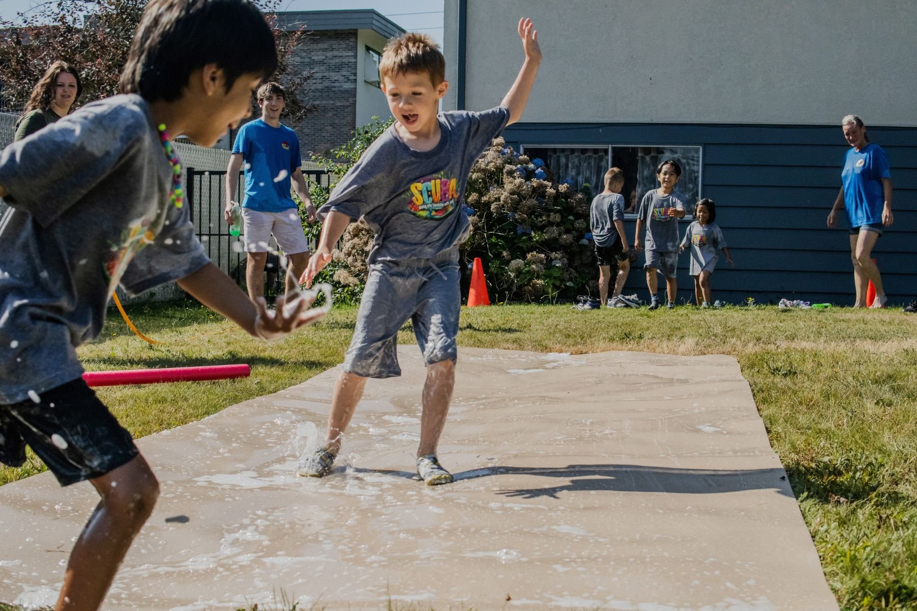 Kids playing on slip 'n slide outside on a sunny day, with some adults in the background.