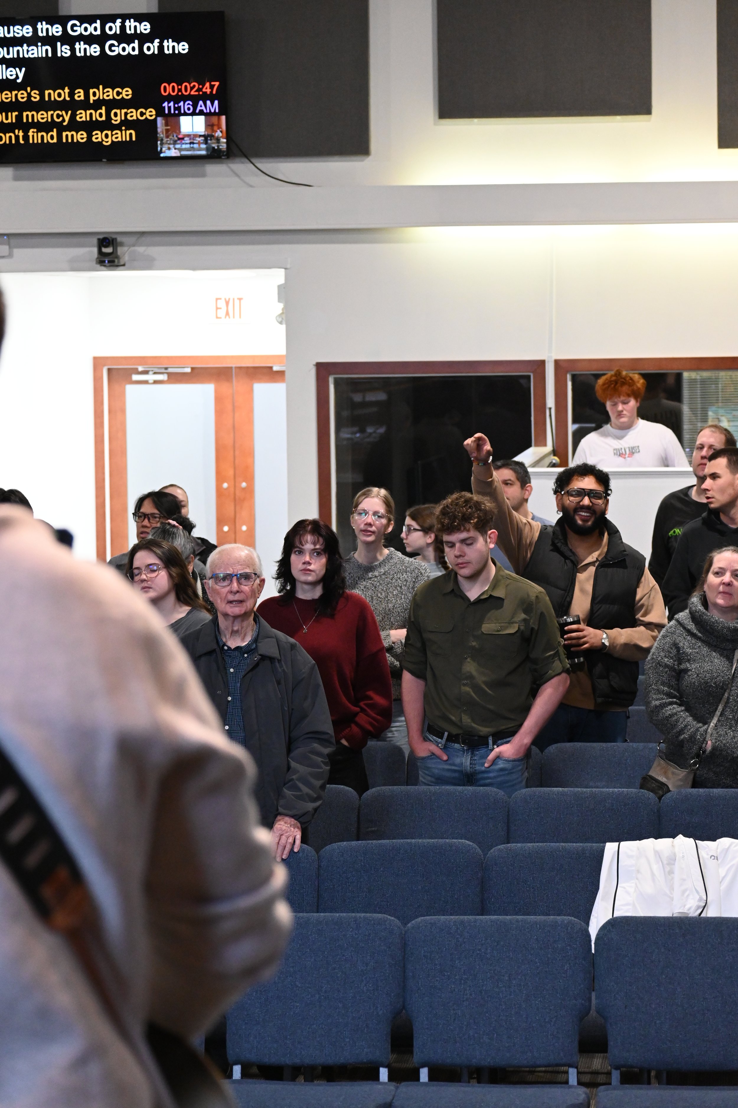 A group of people standing in a room, some with hands in pockets, others looking forward or to the side, with a large digital screen overhead displaying text and a clock, and blue chairs in the foreground. A white jacket is draped over one of the chairs.