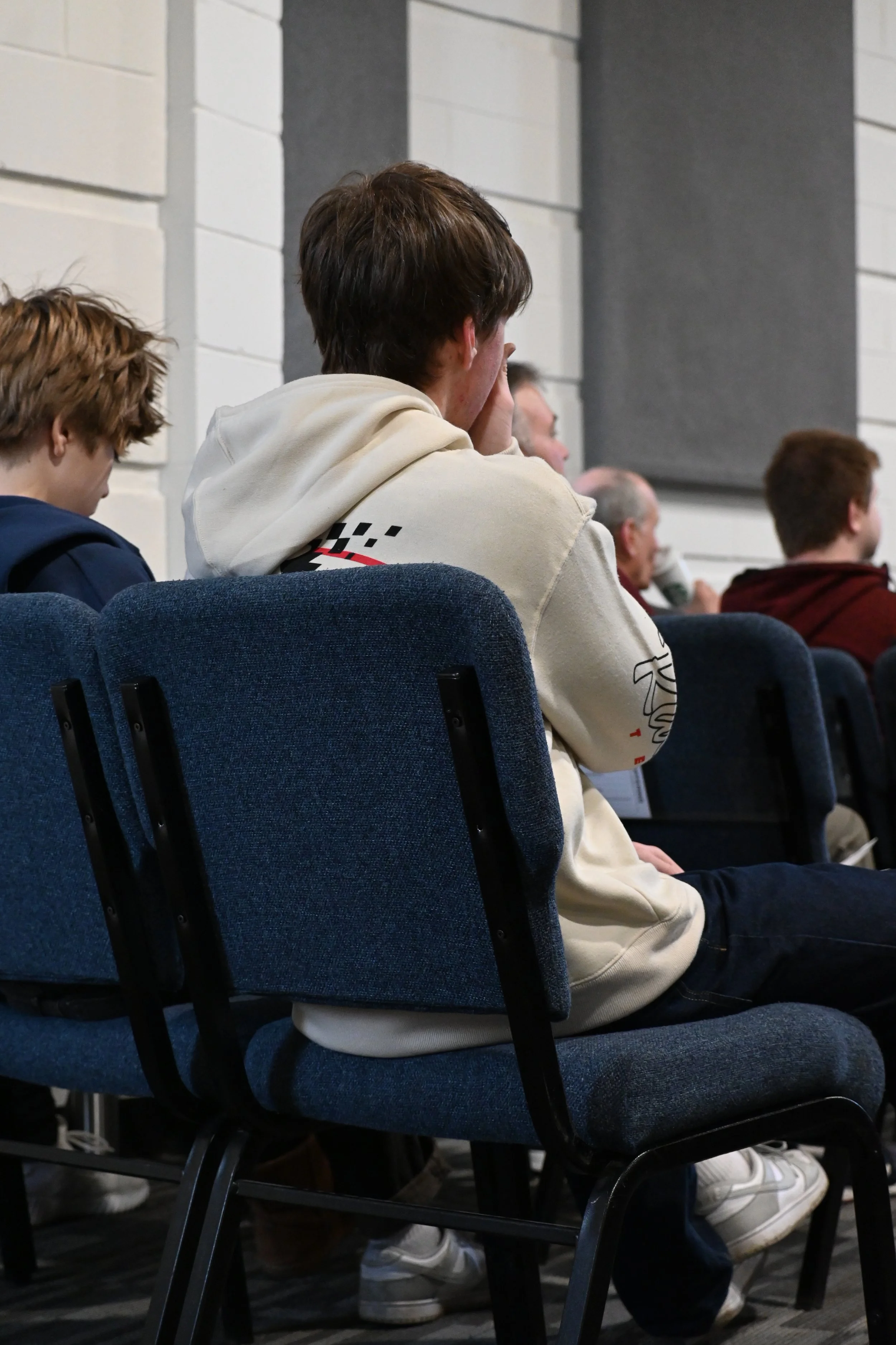 A young man with brown hair wearing a white hoodie, sitting on a blue chair, listening attentively at a church service or event with other attendees.