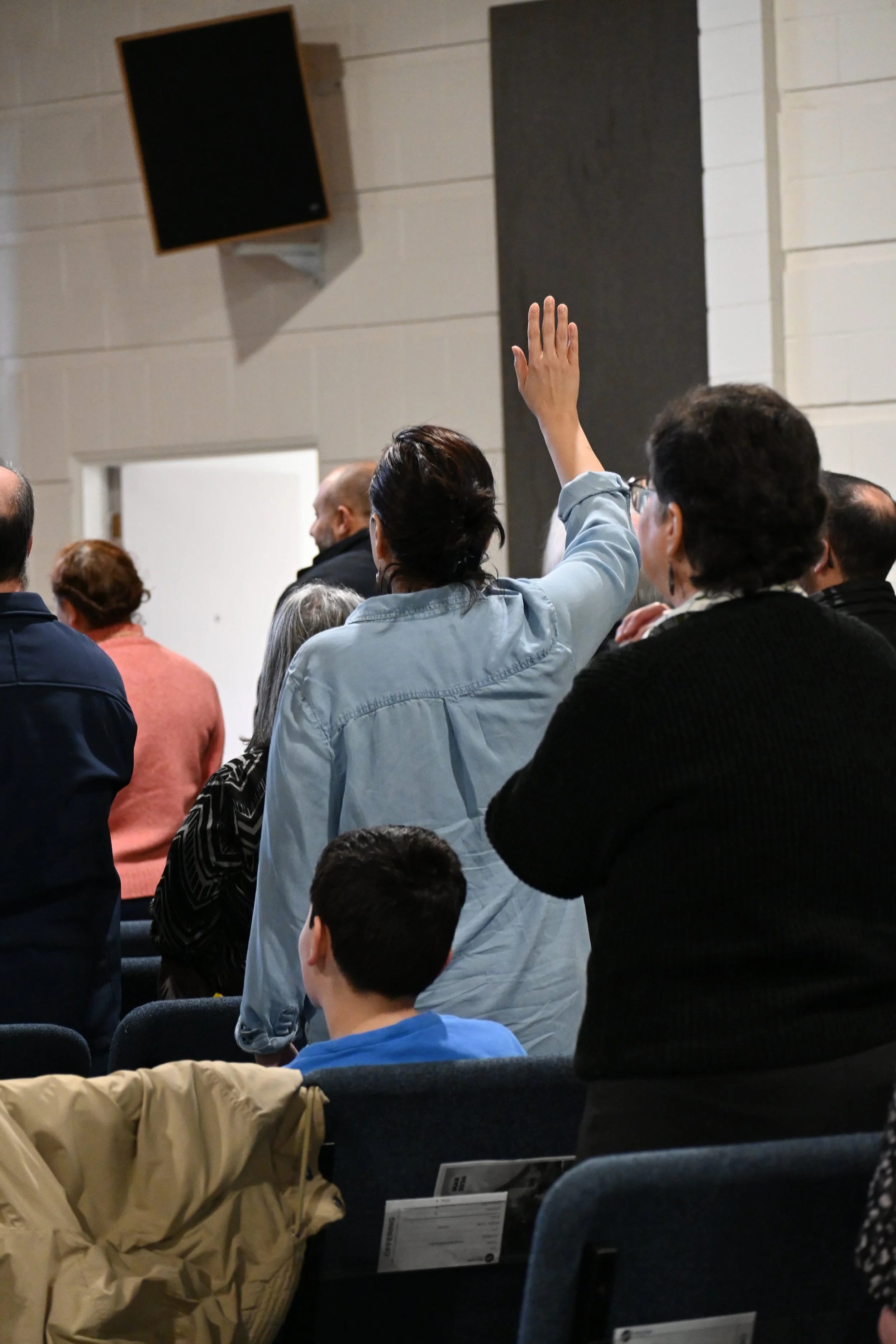 A woman with dark hair and wearing a light blue shirt is raising her hand in a crowded indoor meeting or conference.