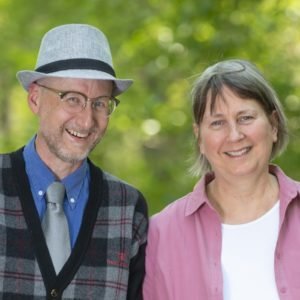 A smiling man wearing a grey fedora and glasses, and a woman with short hair, outdoors with green foliage in the background.
