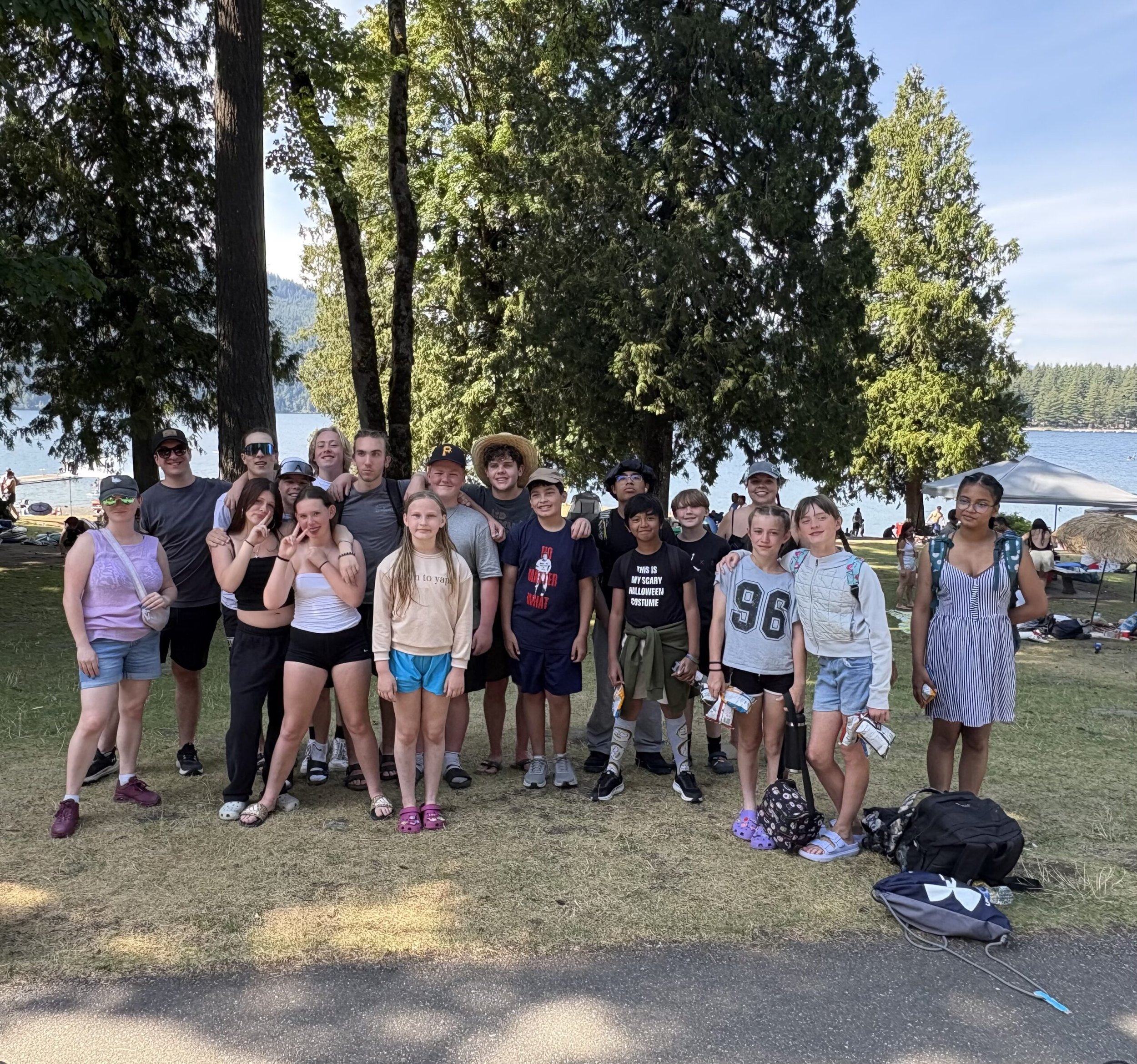 A group of children and teenagers posing outdoors in front of trees with a lake and forest in the background, some holding snacks.