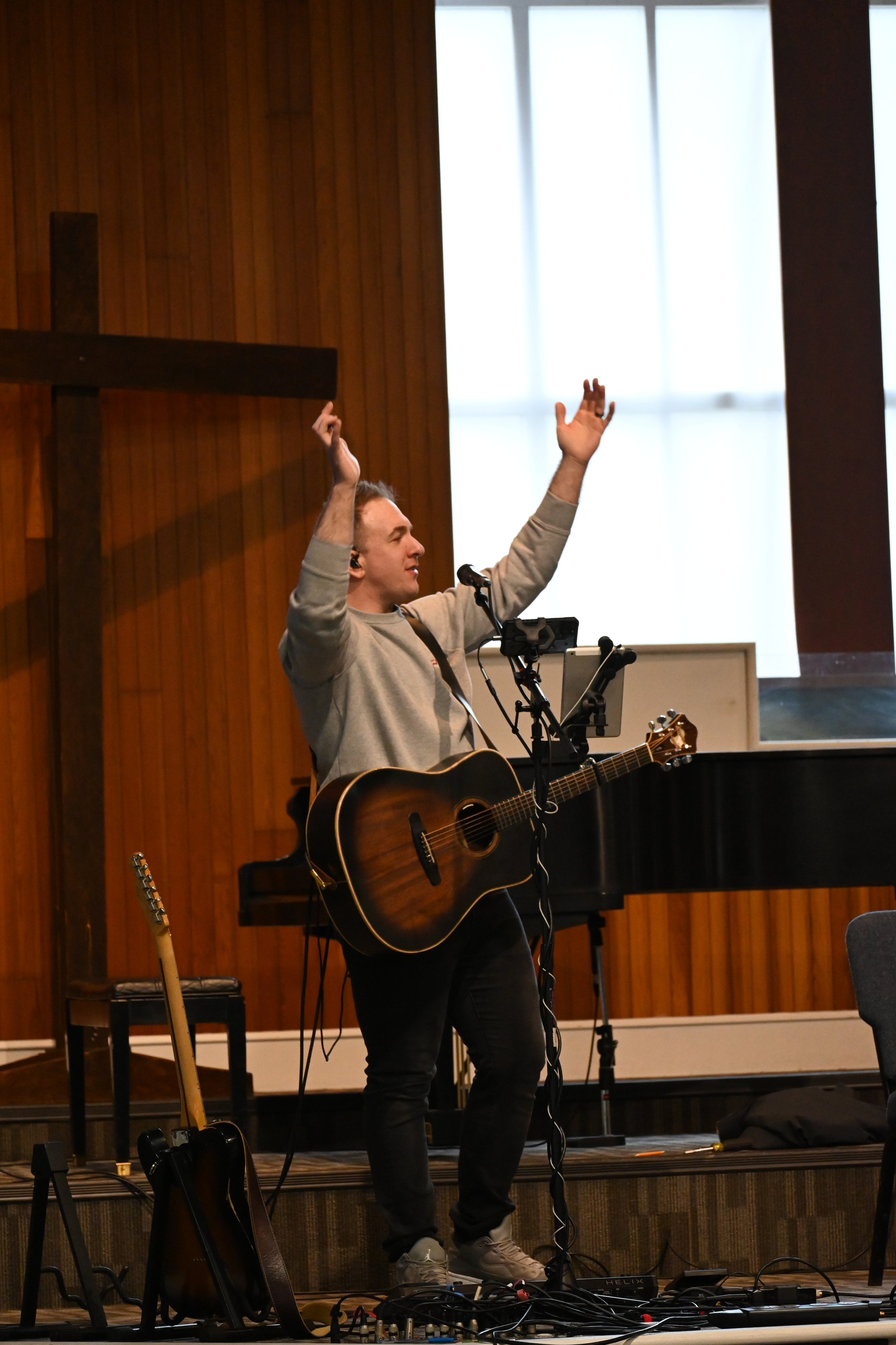 A man playing guitar and singing on stage with his arms raised, in a church or performance hall, with a large wooden cross and a piano in the background.
