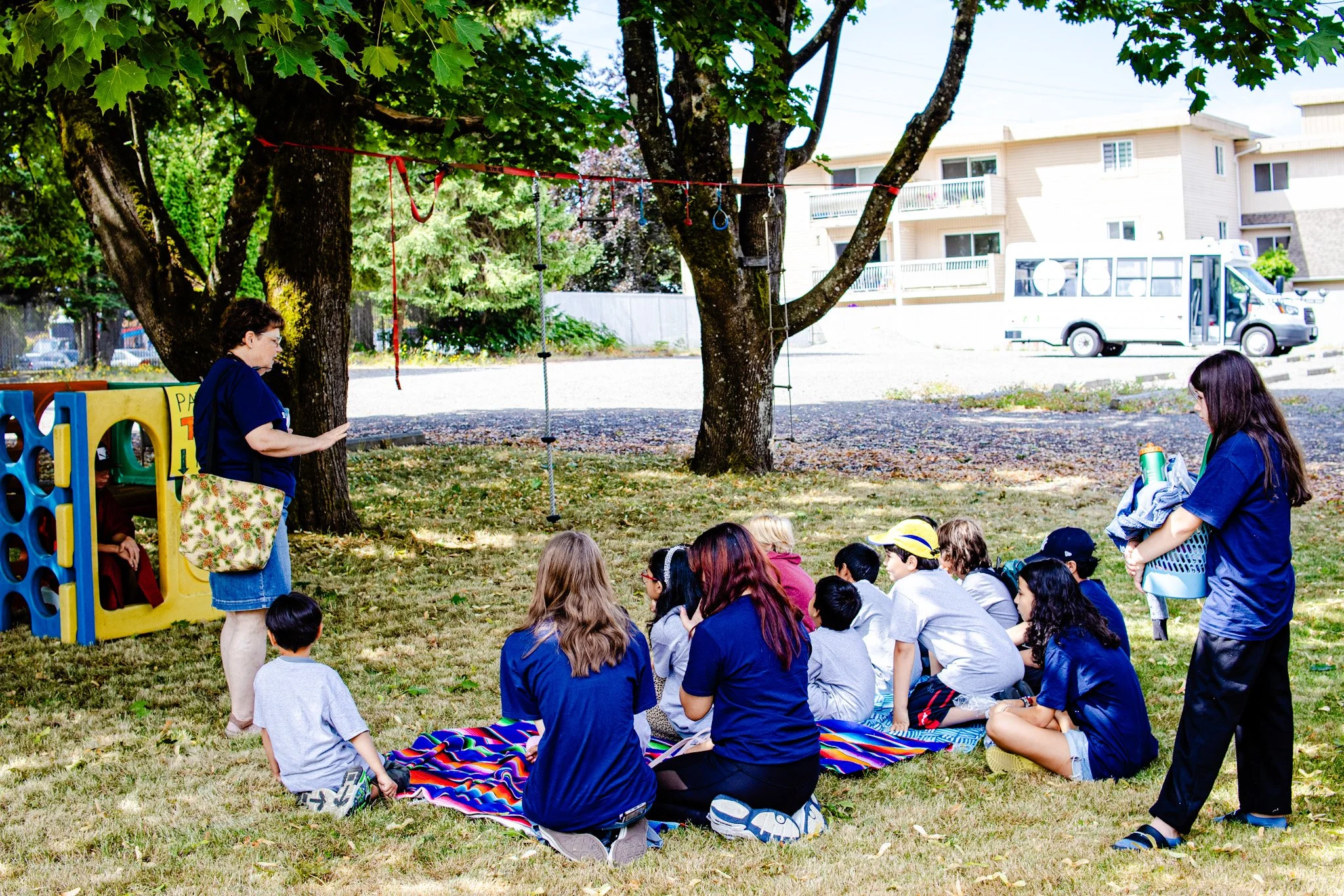 Group of children sitting on a colorful blanket under a tree during an outdoor activity, with two women supervising, one standing and the other carrying supplies, in a park-like setting.