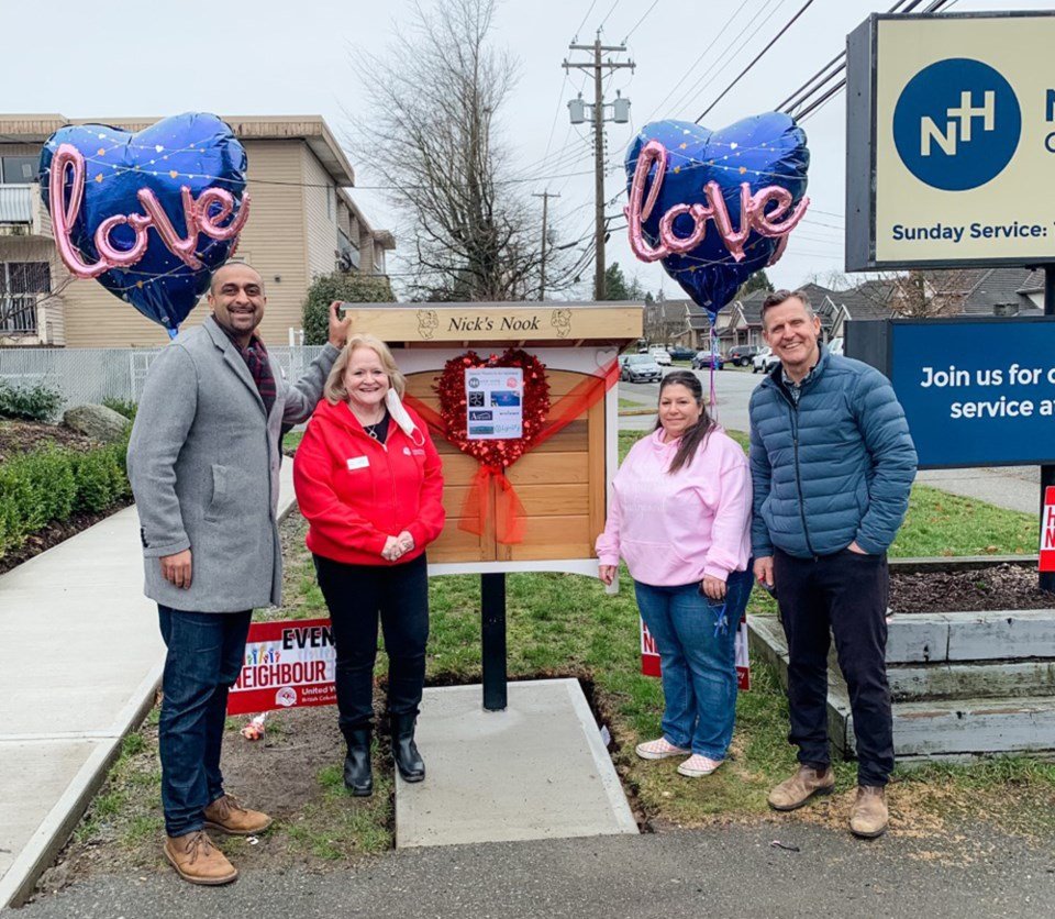 Four people stand in front of a small wooden structure with a sign that says "Nick's Nook". There are two blue heart-shaped balloons with pink "love" written on them, and a red heart decoration on the structure. The scene appears to be a community or charity event.