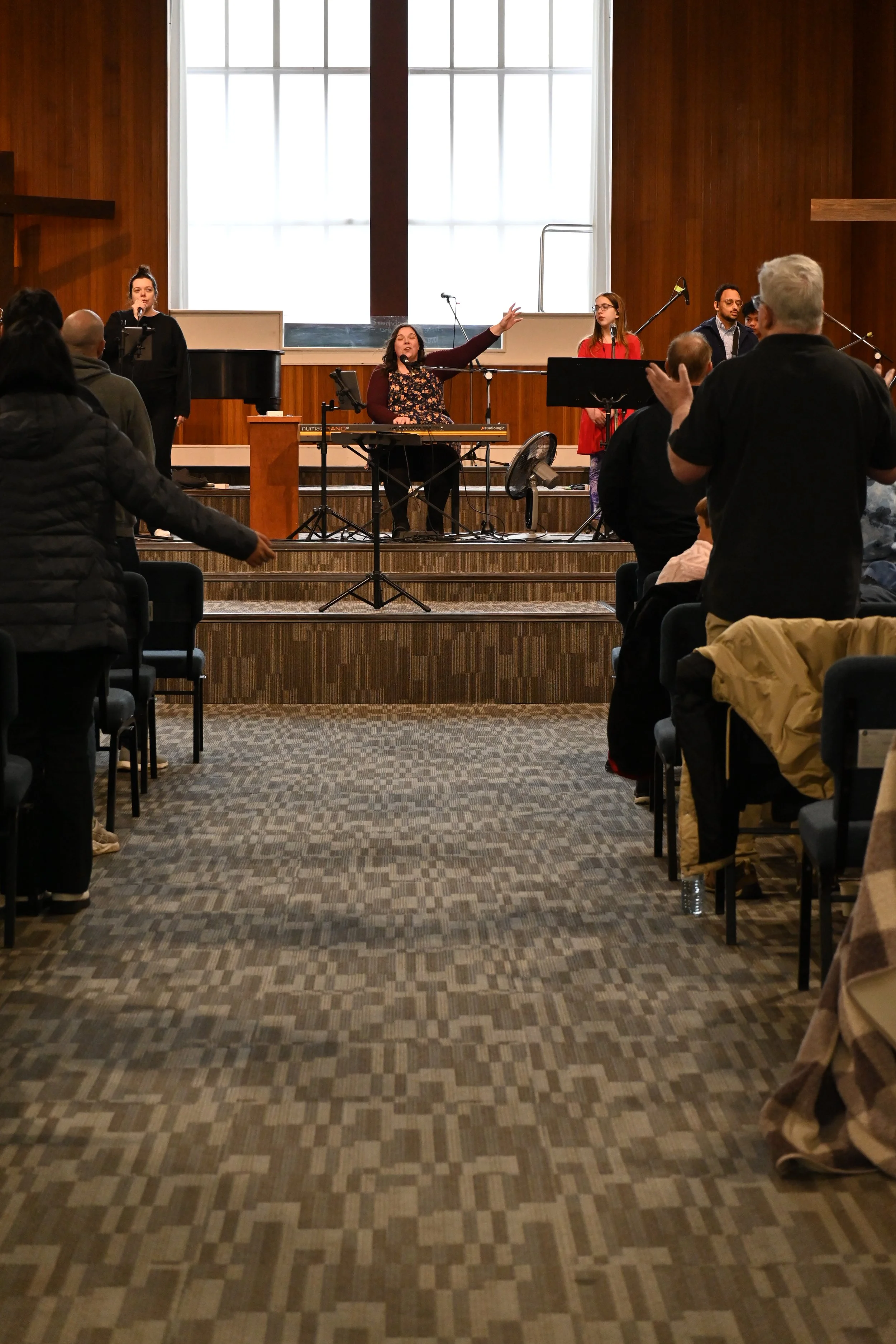 A musical performance taking place on a stage in a church, with a woman singing into a microphone on the left, four musicians with instruments and microphones on the right, and several congregant members standing and clapping.