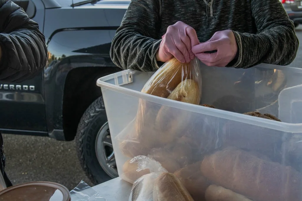 Person's hands holding a plastic bag of baked goods over a clear plastic container filled with more baked goods, with a black Toyota Tacoma truck in the background.