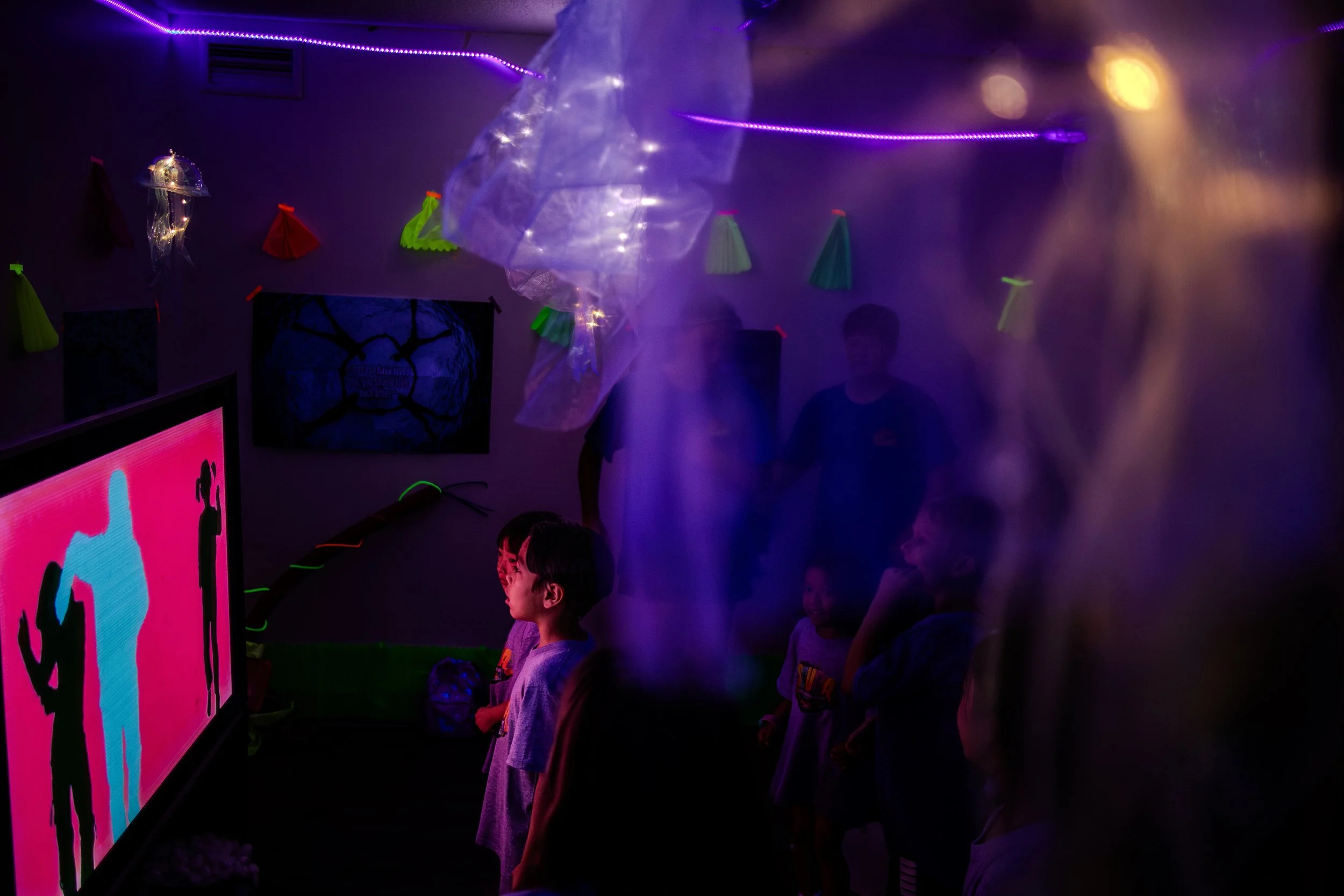 Children and adults watching a glow dance on a screen in a dark room decorated with paper umbrellas and neon lights.