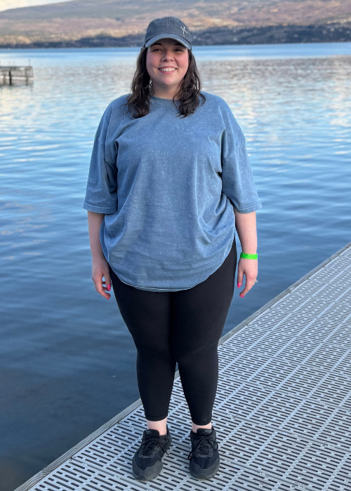 Young woman smiling on a dock by a lake, wearing a blue t-shirt, black leggings, a cap, and sneakers.
