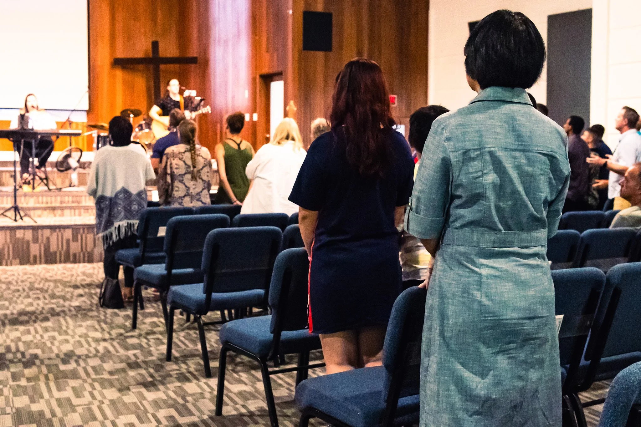 People attending a church service or event, standing and facing a stage with a cross, where musicians are performing, in a church auditorium.