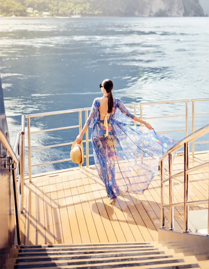 A woman in a flowing blue dress walking down the aft deck of a luxury yacht at sunset, overlooking calm coastal waters.