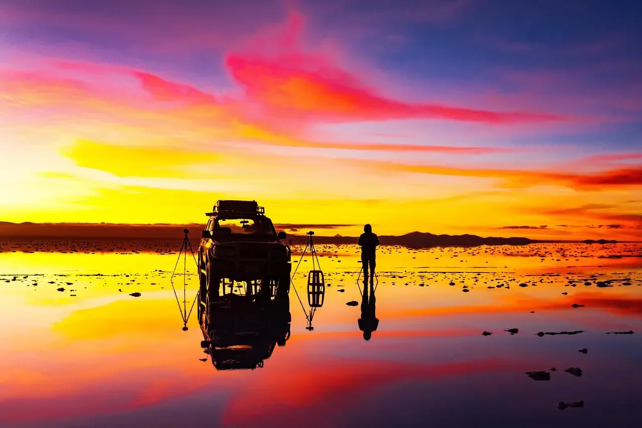 The silhouette of a traveler and a 4x4 expedition vehicle reflected perfectly in the mirror-like surface of the Salar de Uyuni during a vibrant sunset.