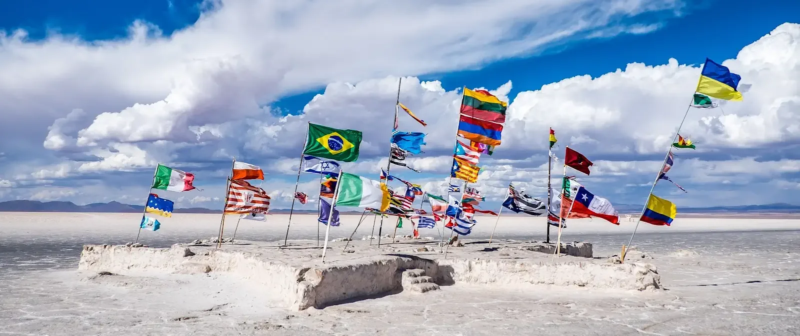 A collection of international flags planted on a salt-block platform across the vast expanse of the Salar de Uyuni in Bolivia.
