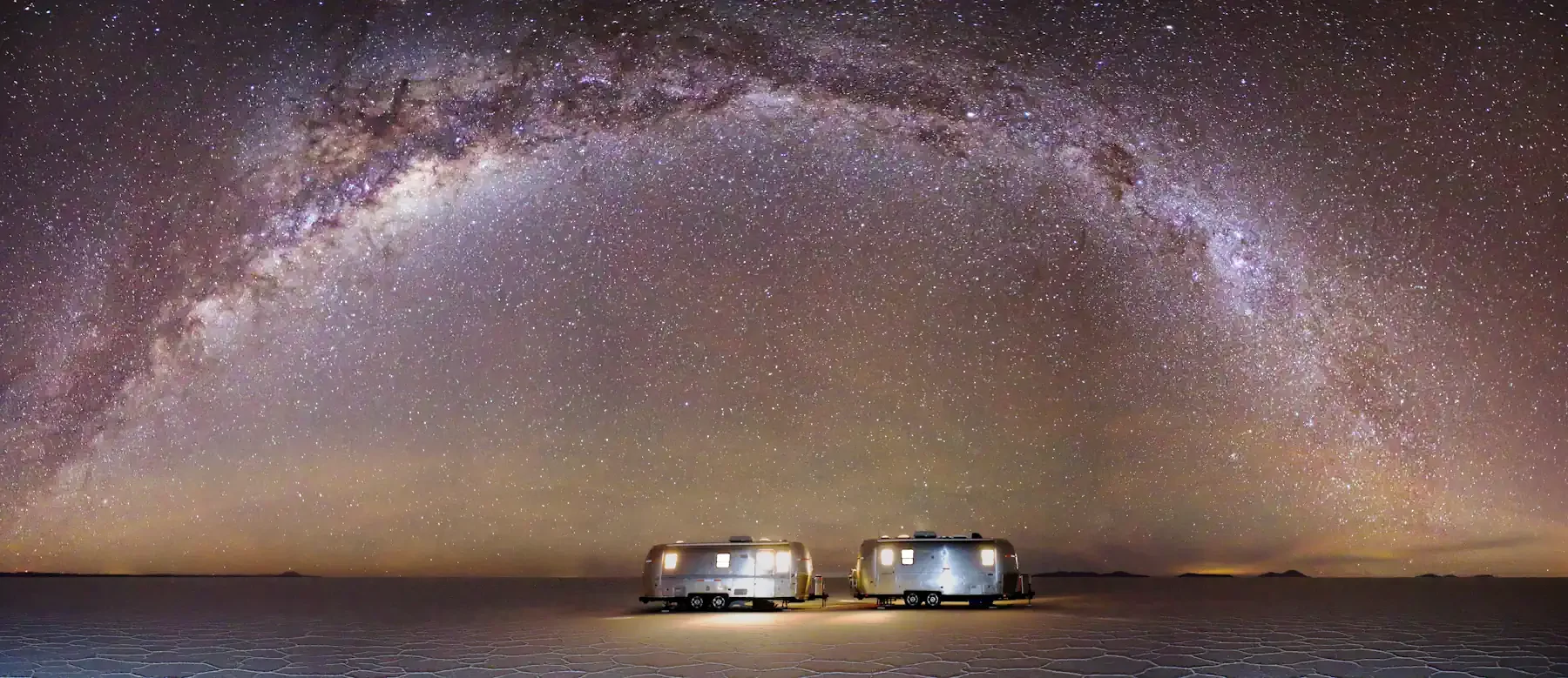 Two private Airstream campers illuminated beneath the Milky Way galaxy on the expansive Salar de Uyuni salt flats in Bolivia.