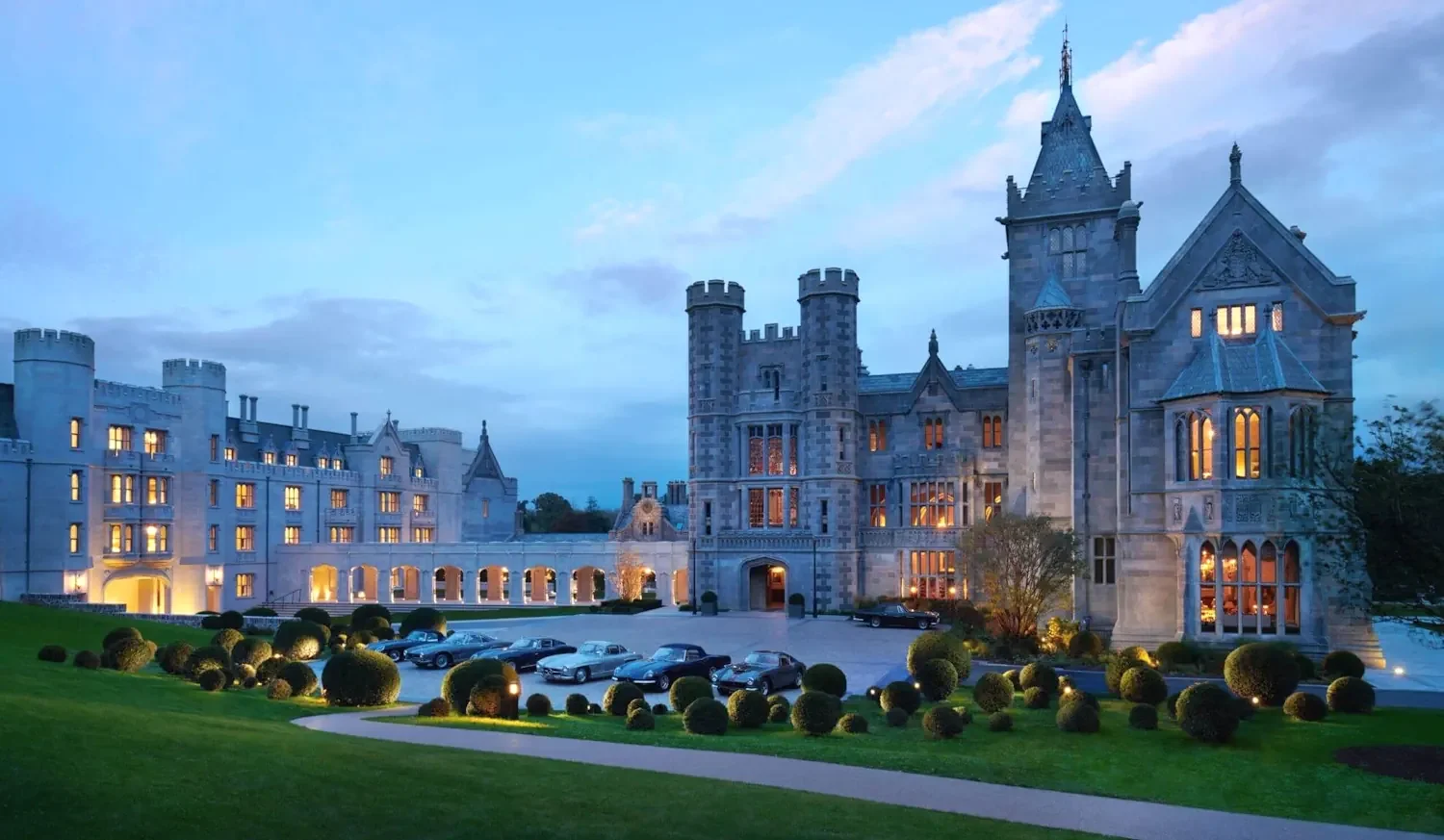 An evening view of the illuminated Gothic Revival architecture at Adare Manor's grand entrance, featuring a collection of vintage luxury cars in the courtyard.