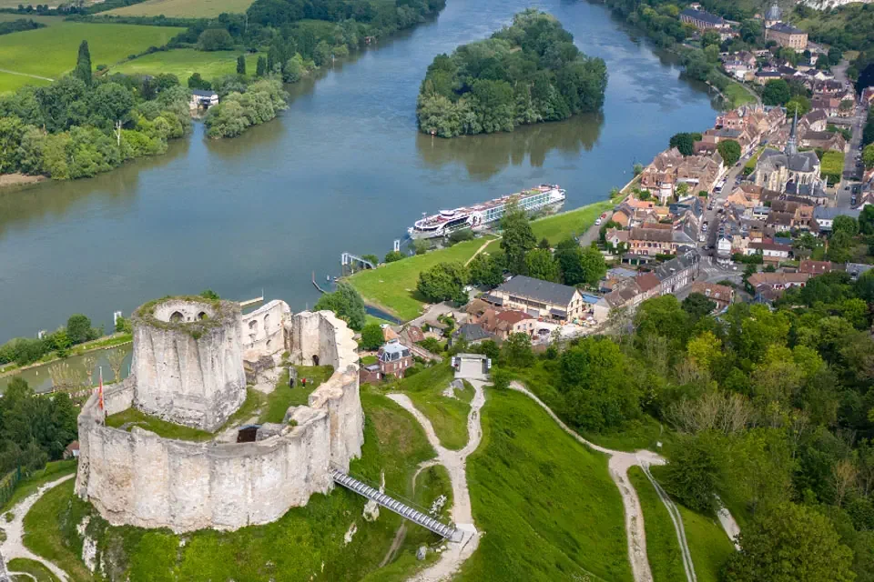 An aerial view of a Tauck luxury riverboat docked on the Seine River in Les Andelys, France, showcasing the scenic landscapes of European river cruising beneath the historic Château Gaillard.