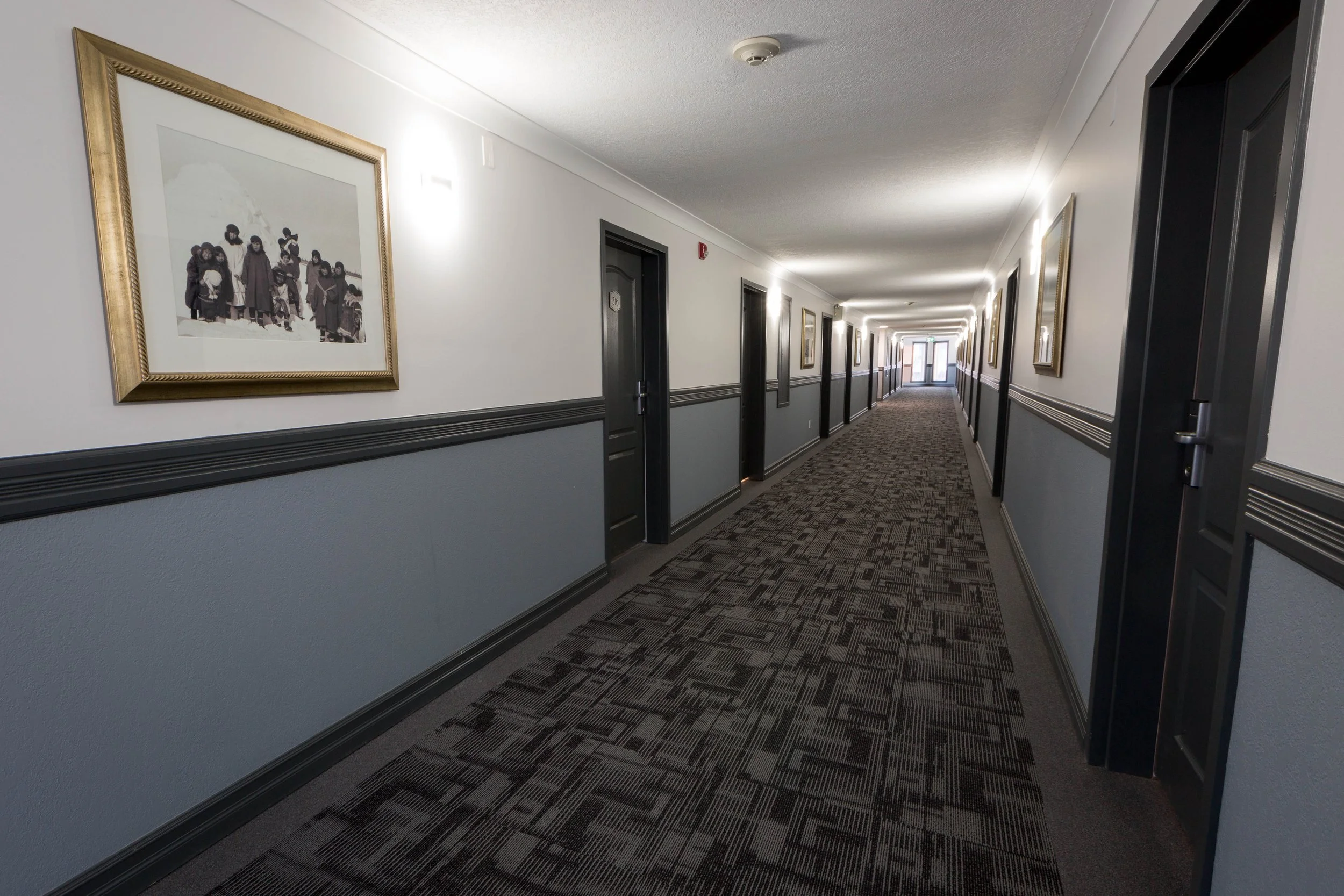 An indoor hallway in a hotel or apartment building with framed artwork on the walls, multiple closed doors, and patterned carpet flooring.