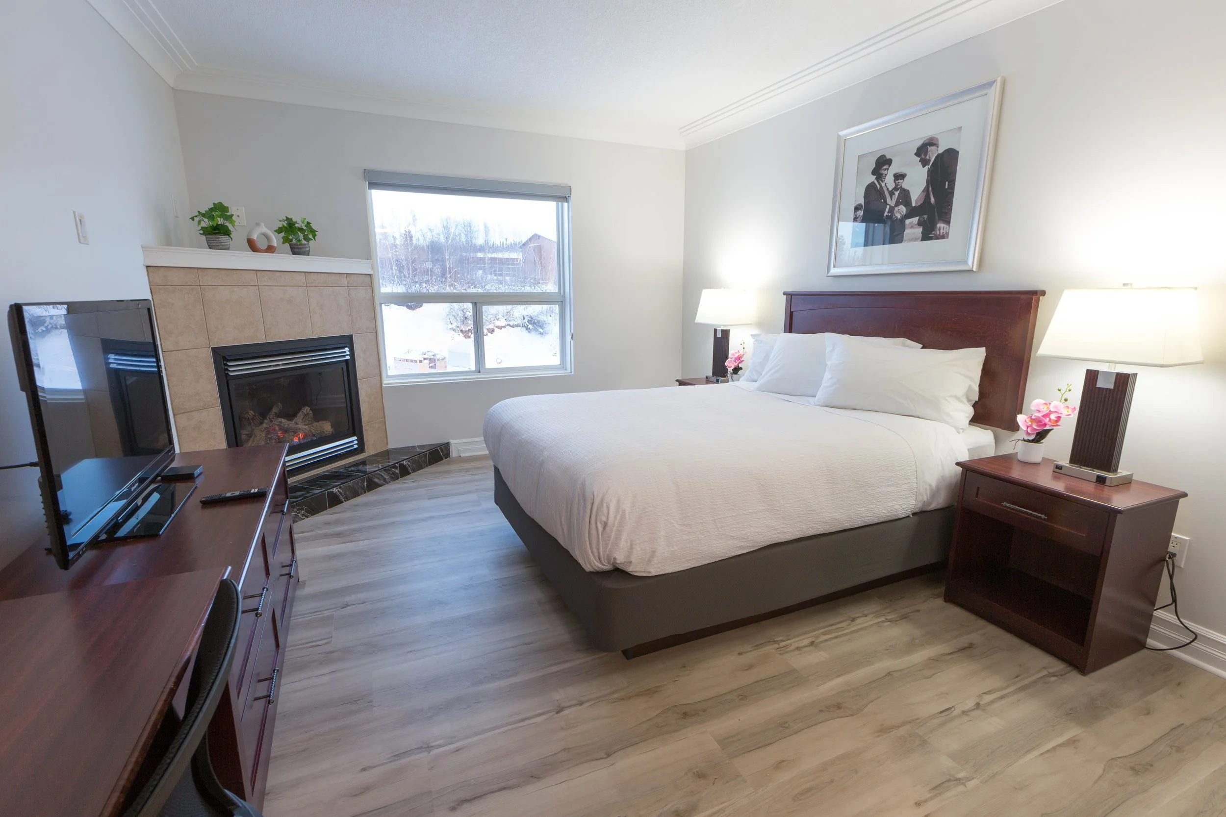 A hotel room with a bed, nightstands, lamps, a fireplace, a television, and artwork on the wall, with a window showing a snowy landscape outside.