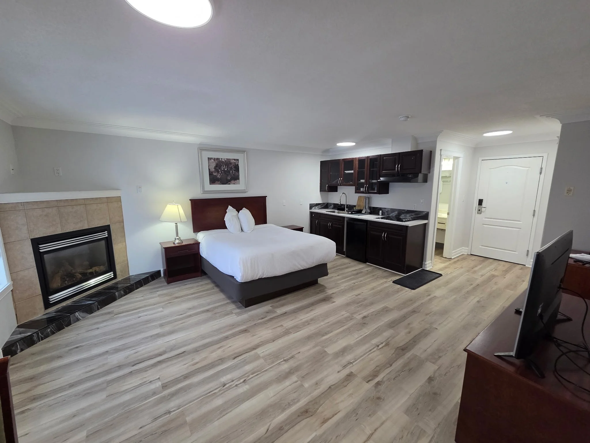 A studio apartment with a bed, small kitchen, fireplace, and wooden flooring.