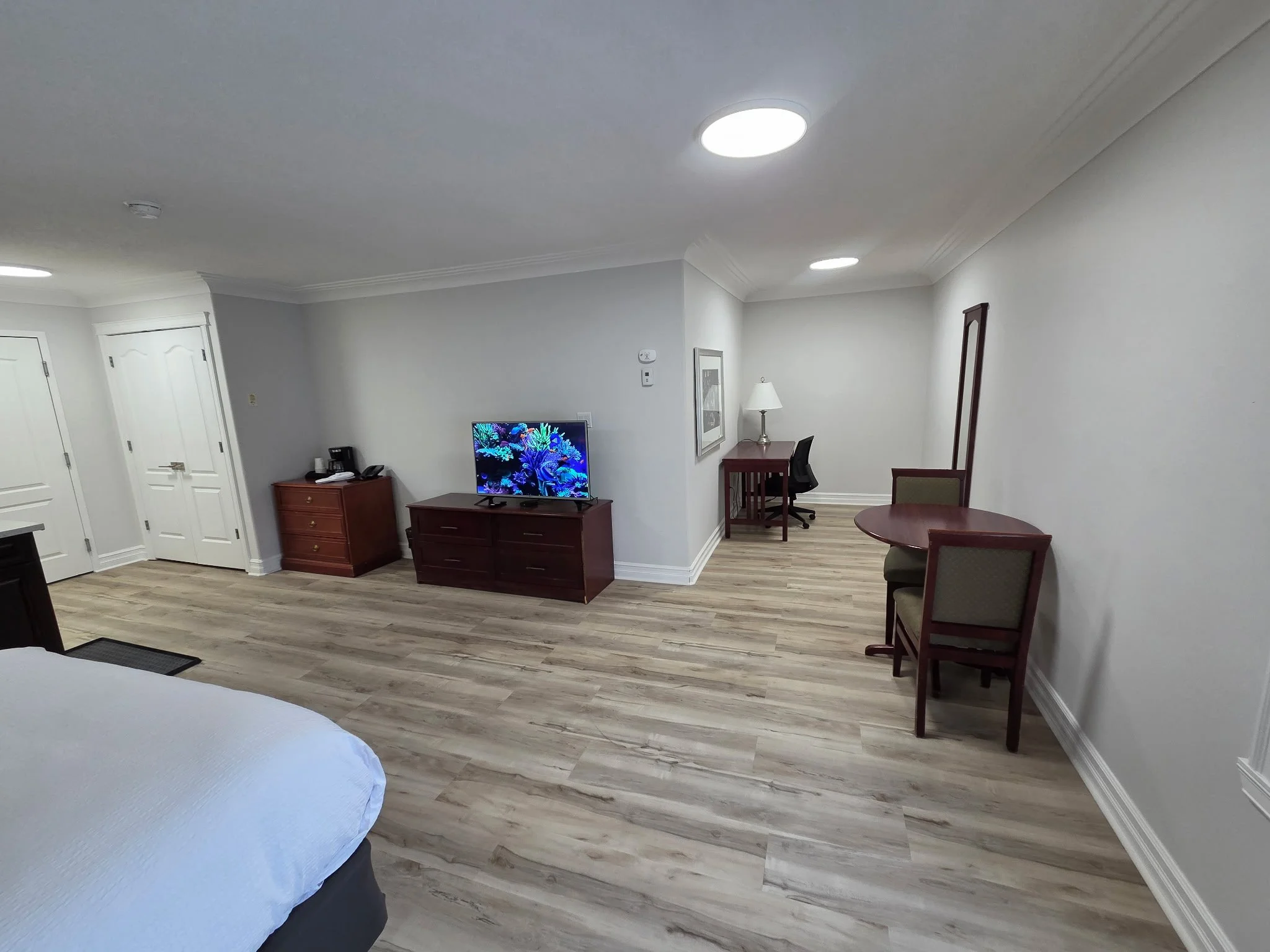 A hotel or apartment room with a wooden floor, a white ceiling with recessed lighting, and white walls. There is a small wooden desk with a lamp, a television, a chair, and some furniture.