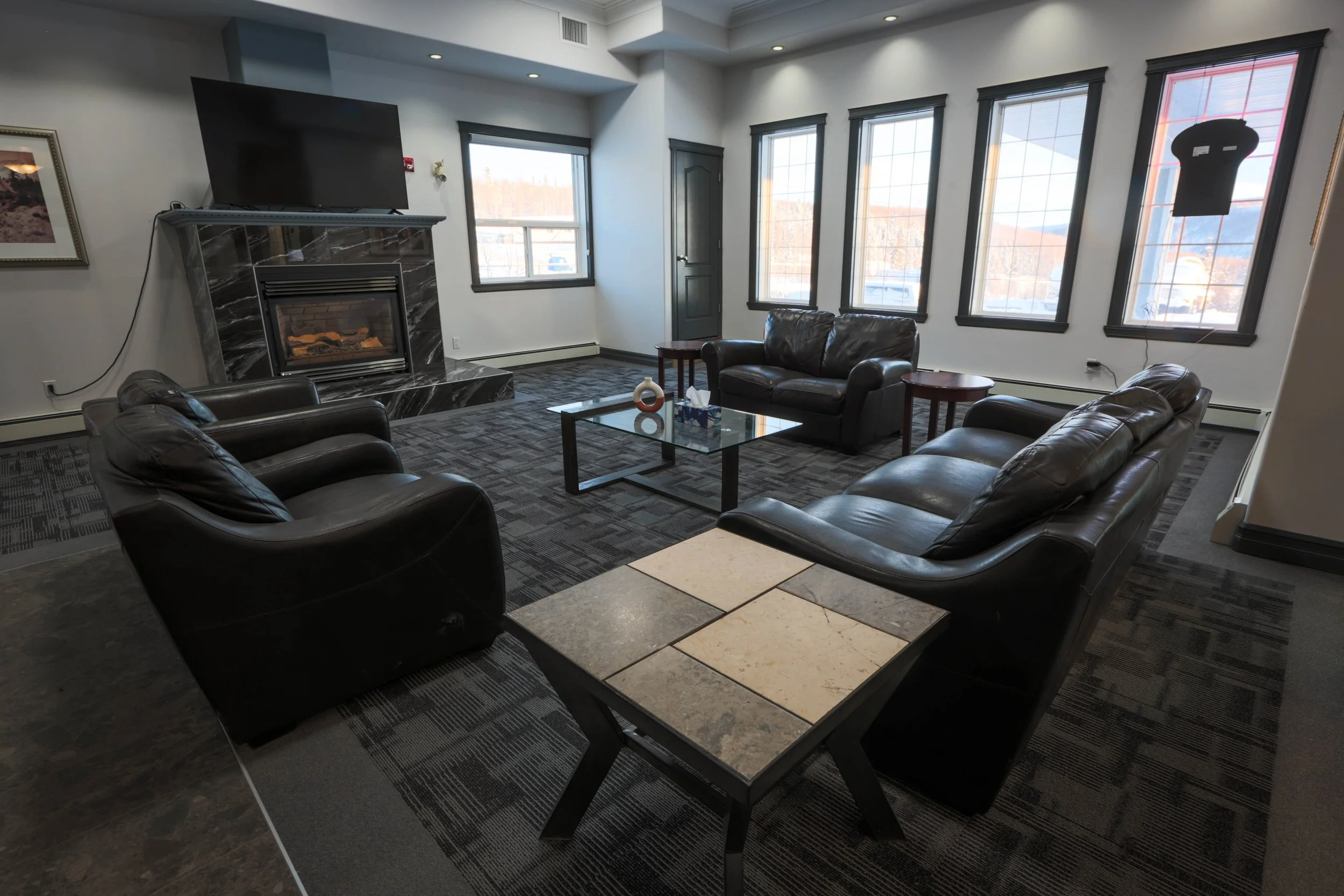 A cozy lounge area with black leather sofas arranged around a coffee table with marble tiles, in front of multiple large windows that let in natural light and offer a view of snow-covered cars and mountains outside. A fireplace with a black and white