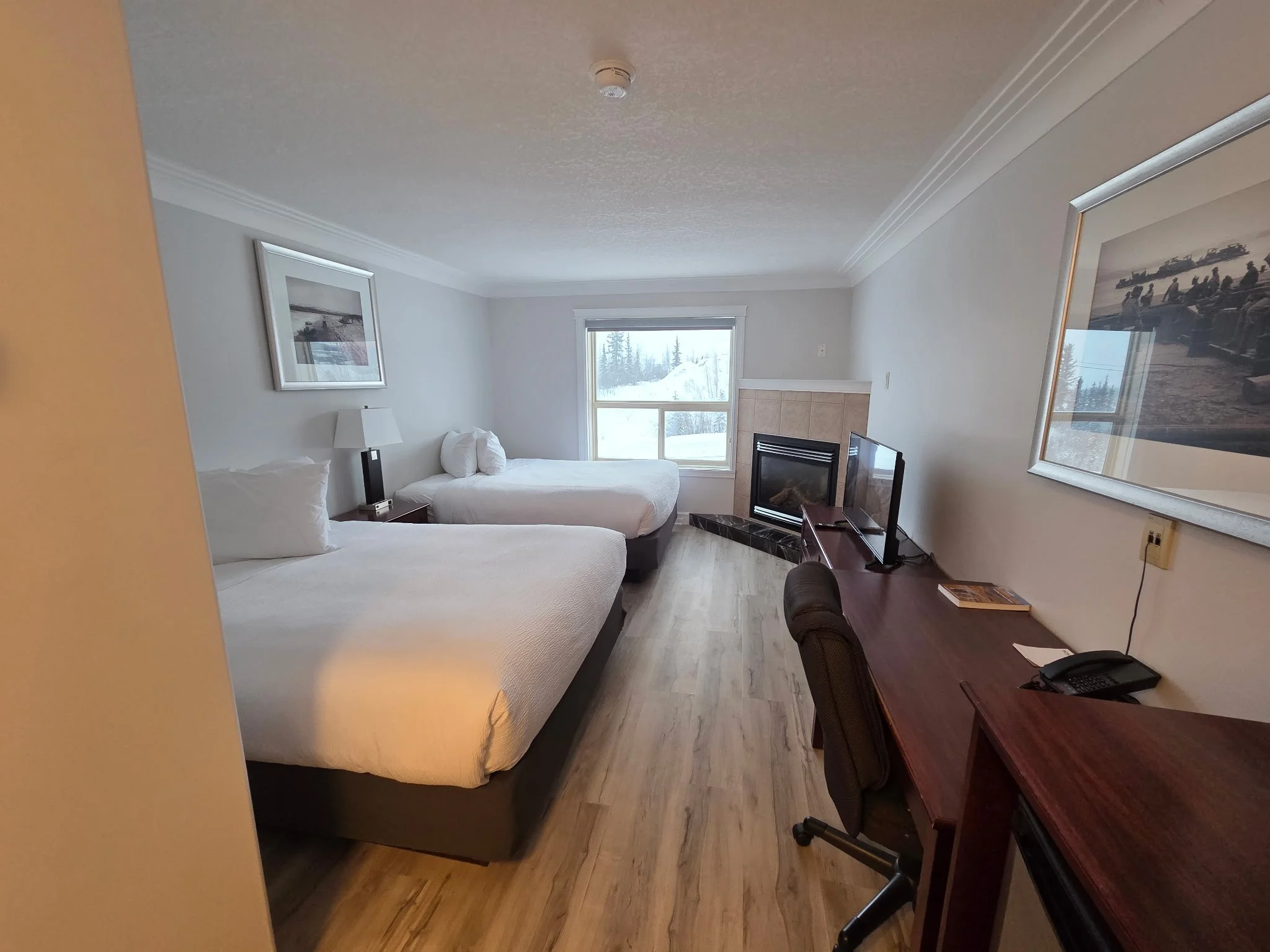 A hotel room with two beds, a window showing snowy landscape and trees, a fireplace, a TV, a desk with a phone, and framed artwork on the walls.