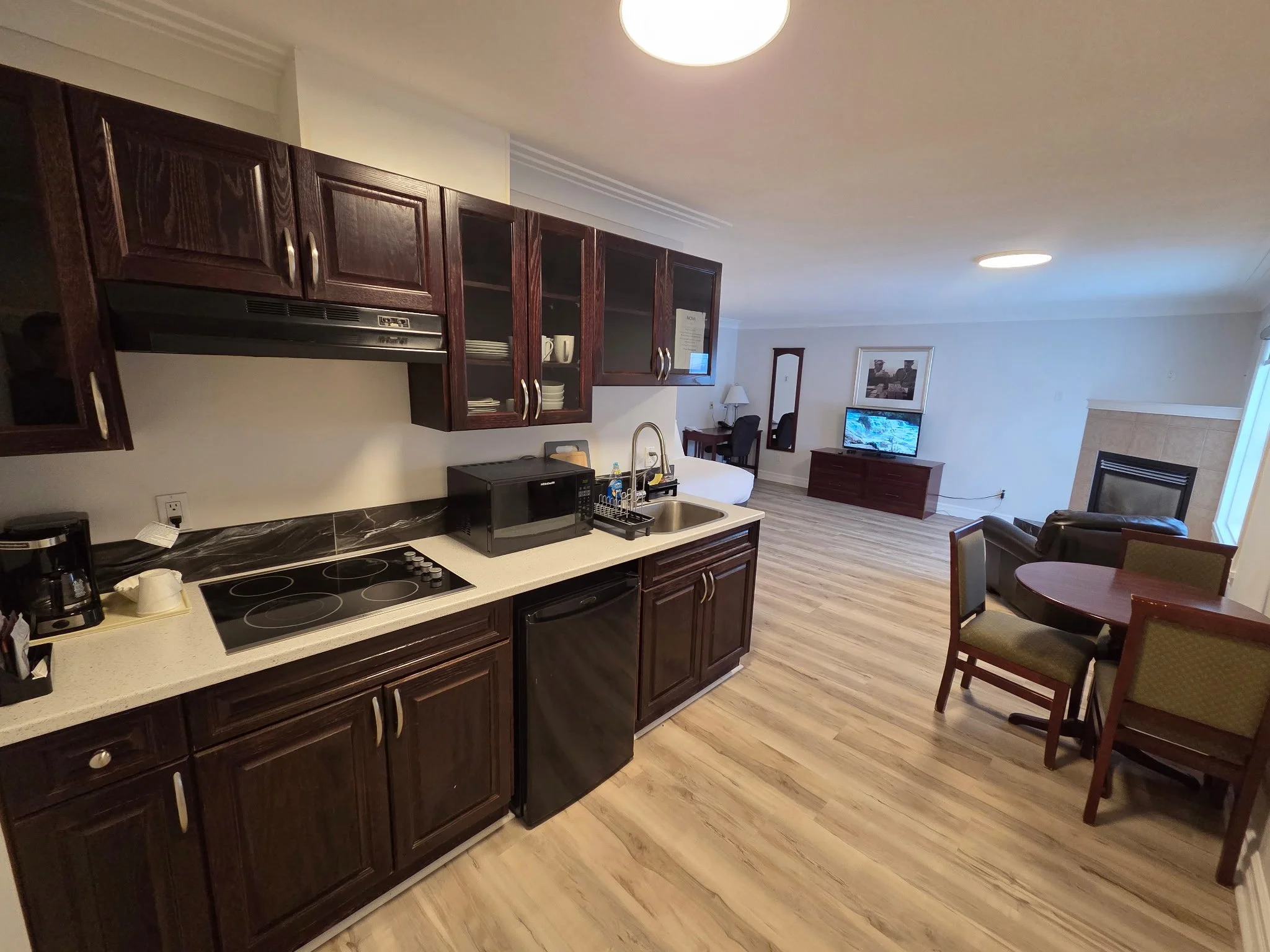 Open-concept kitchen with dark wood cabinets, white countertop, and small appliances, leading into living area with a TV, fireplace, and dining table.