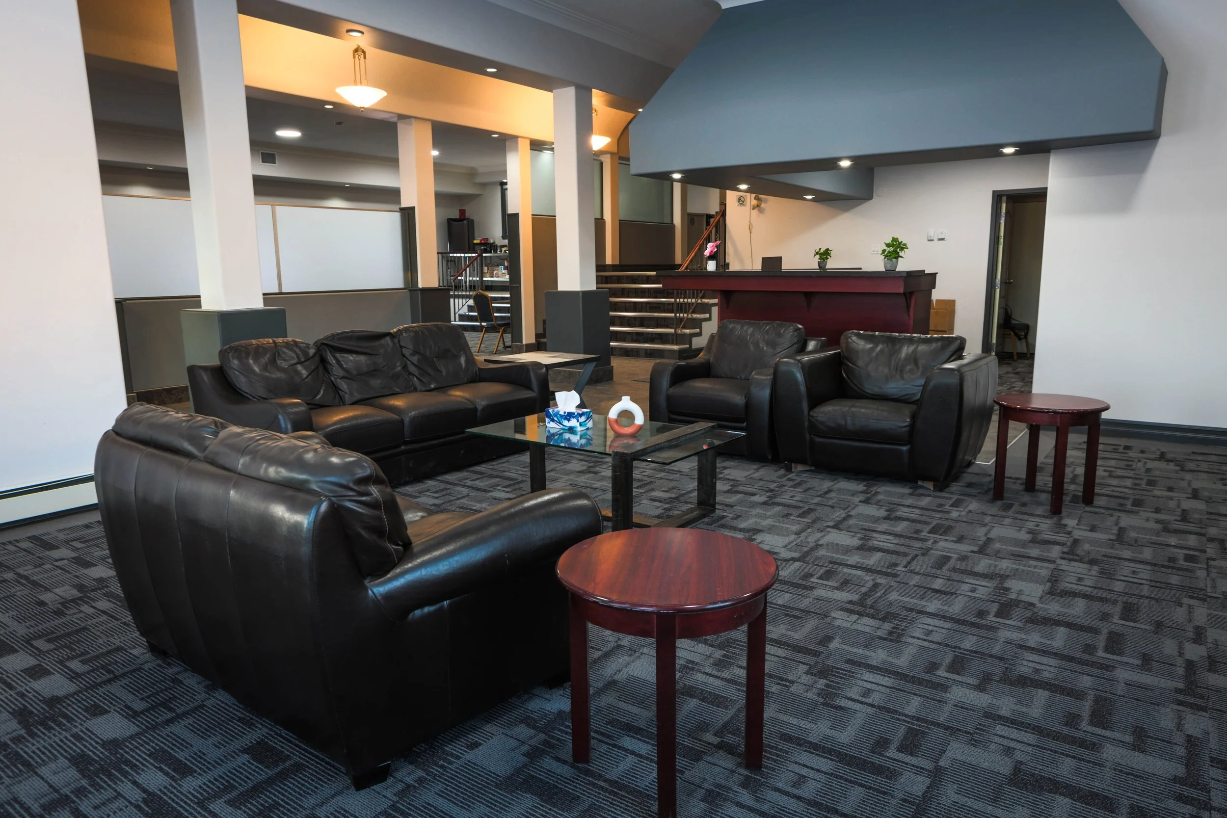 A hotel lobby with black leather sofas, a glass coffee table with decorative items, small red tables, potted plants, stairs with a railing, and ceiling lights, with a carpeted floor.