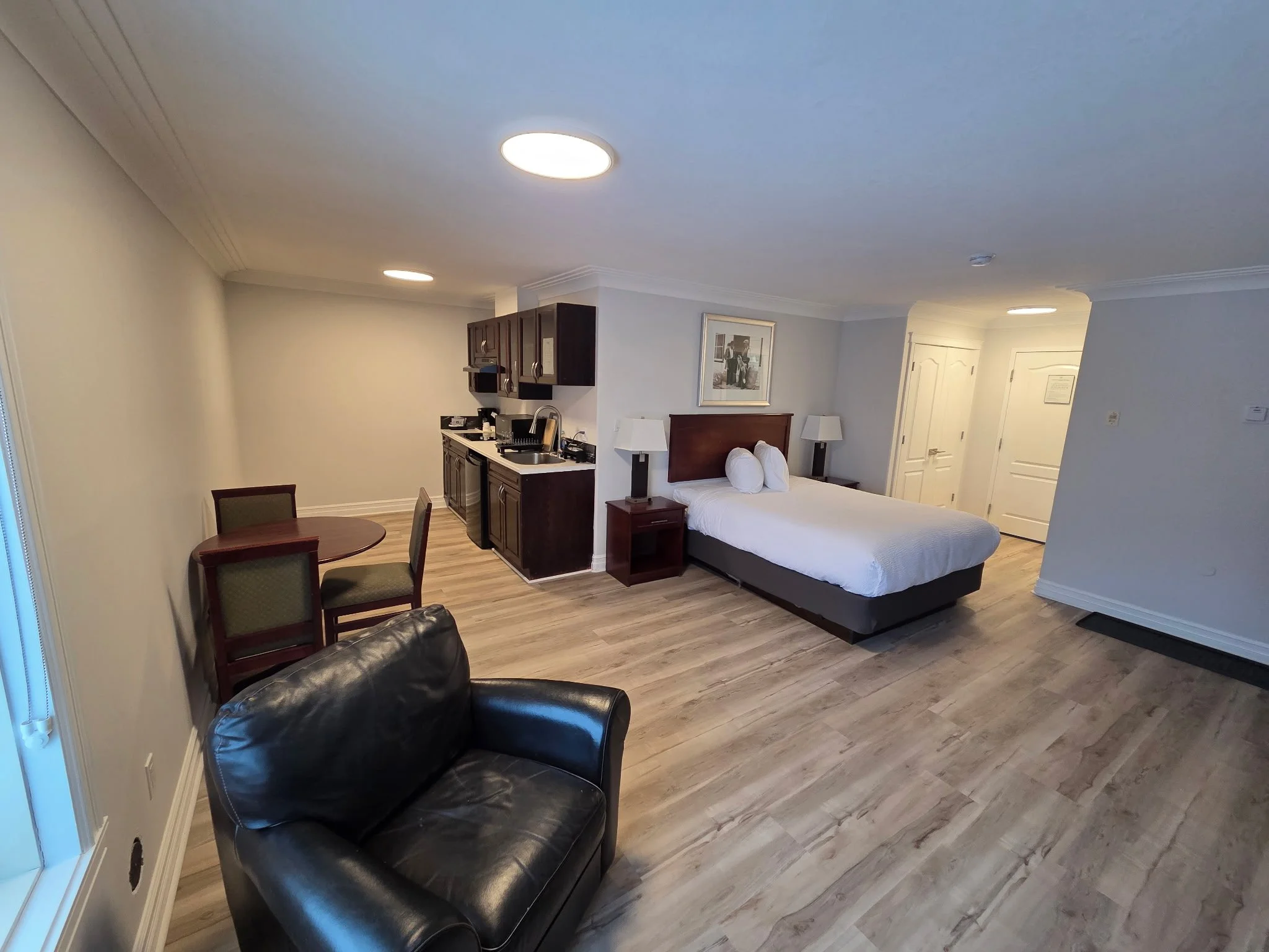 A hotel room with a bed, nightstands, lamps, a painting, a small kitchen area with cabinets, a sink, and a microwave, a round table with chairs, a leather armchair, and a wooden floor.