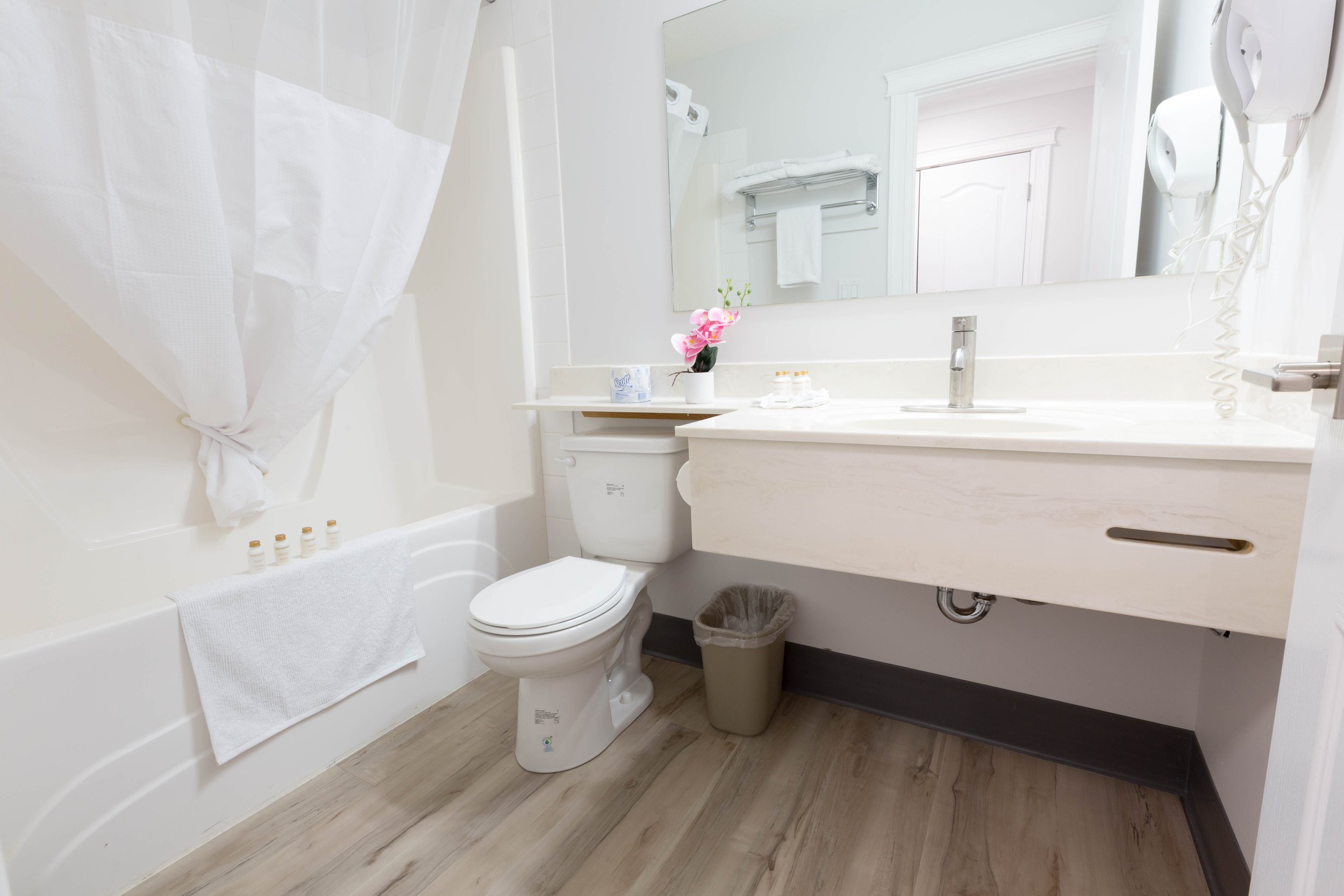 A bathroom with a white bathtub, toilet, and a large mirror above a white sink. There is a small potted pink orchid, toiletries, and a towel on the vanity, and a wall-mounted hairdryer to the side.