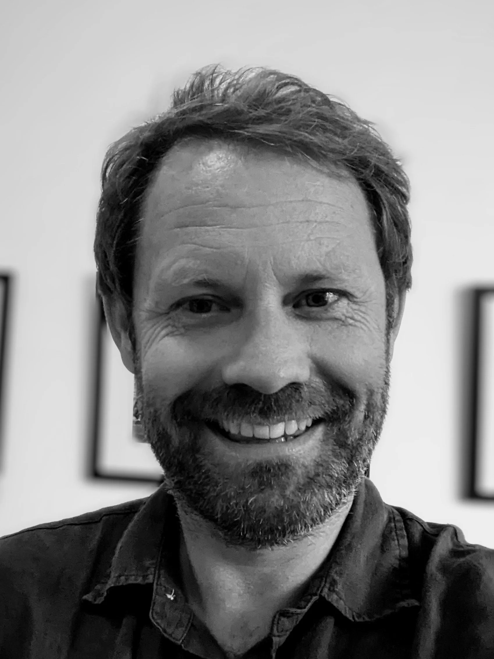 Black and white photo of a man with short, wavy hair and a beard, smiling at the camera, with blurred picture frames in the background.