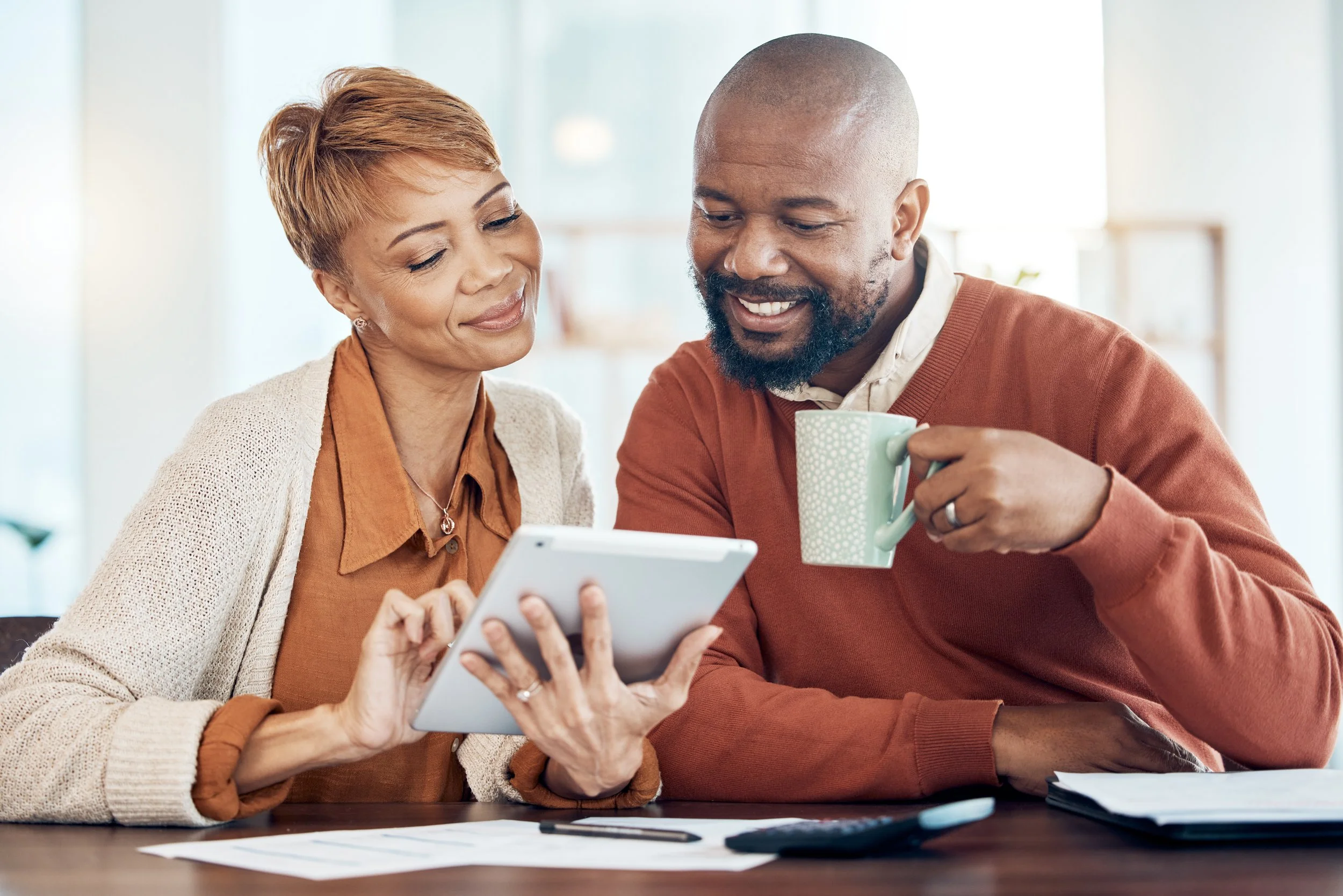 A middle-aged woman and a middle-aged man sitting at a table, looking at a tablet together, with the woman holding the tablet and the man holding a mug