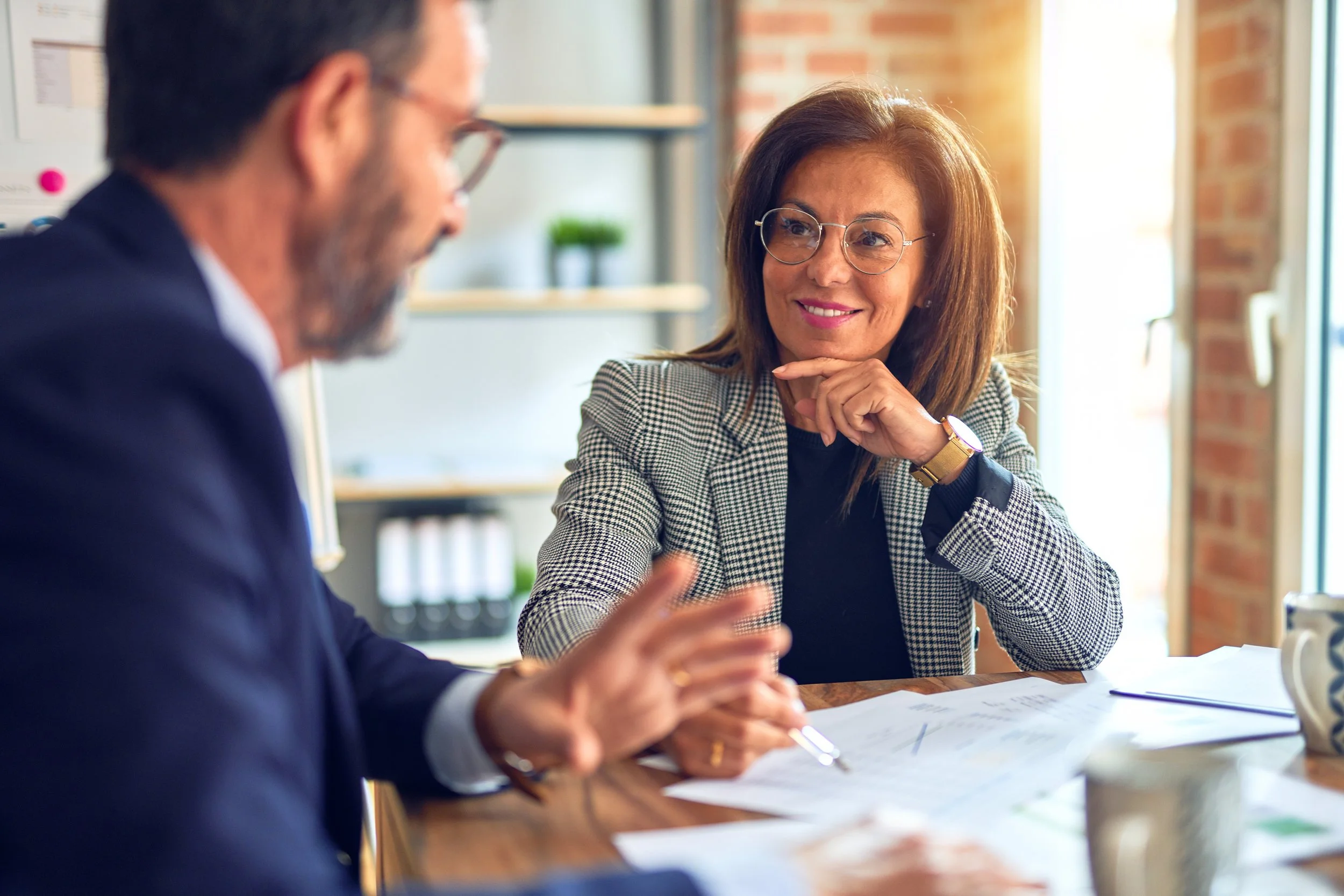 Two professionals having a discussion at a table in a bright office, with a brick wall and large window in the background.