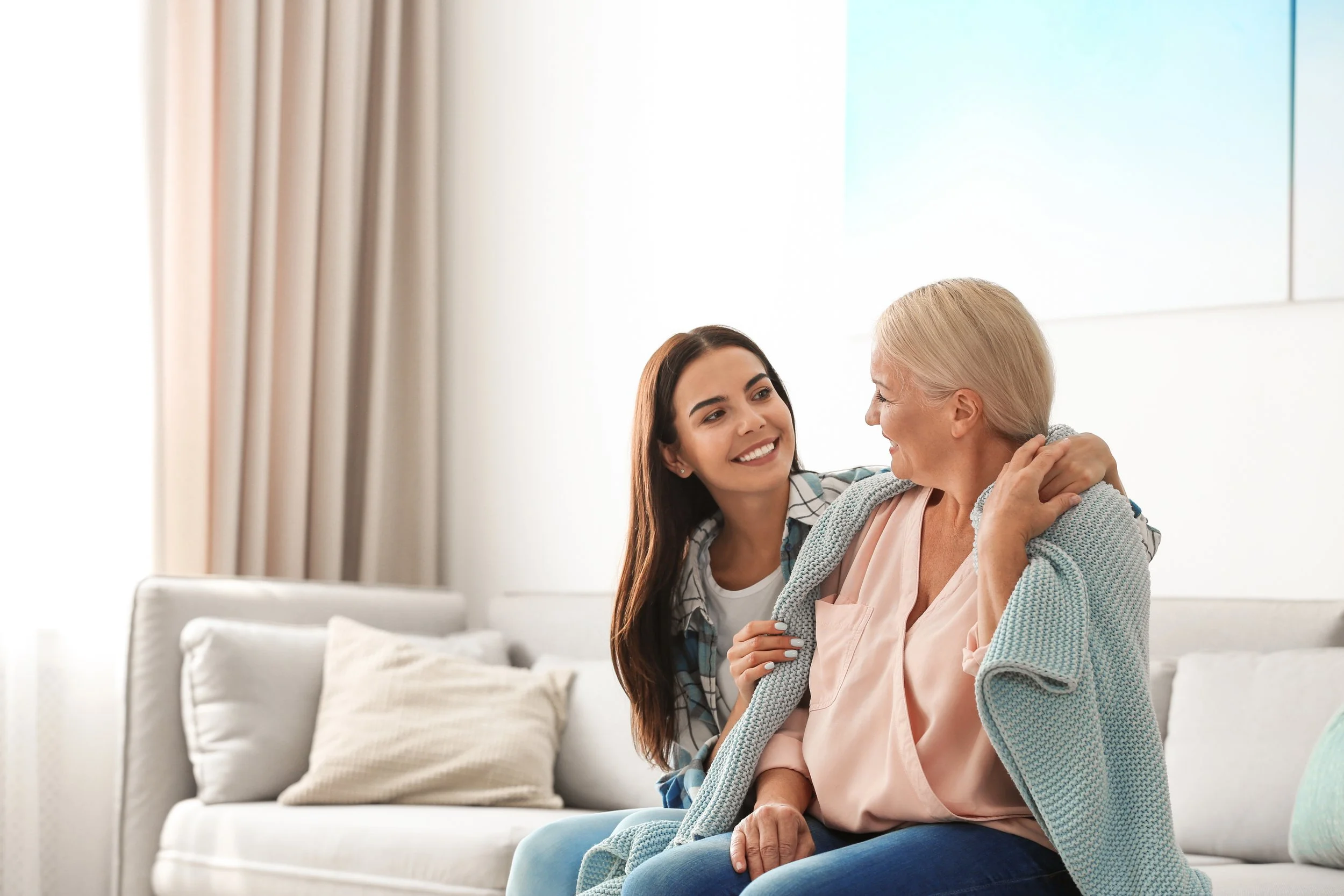 A young woman comforting an elderly woman, sitting together on a sofa in a bright living room.