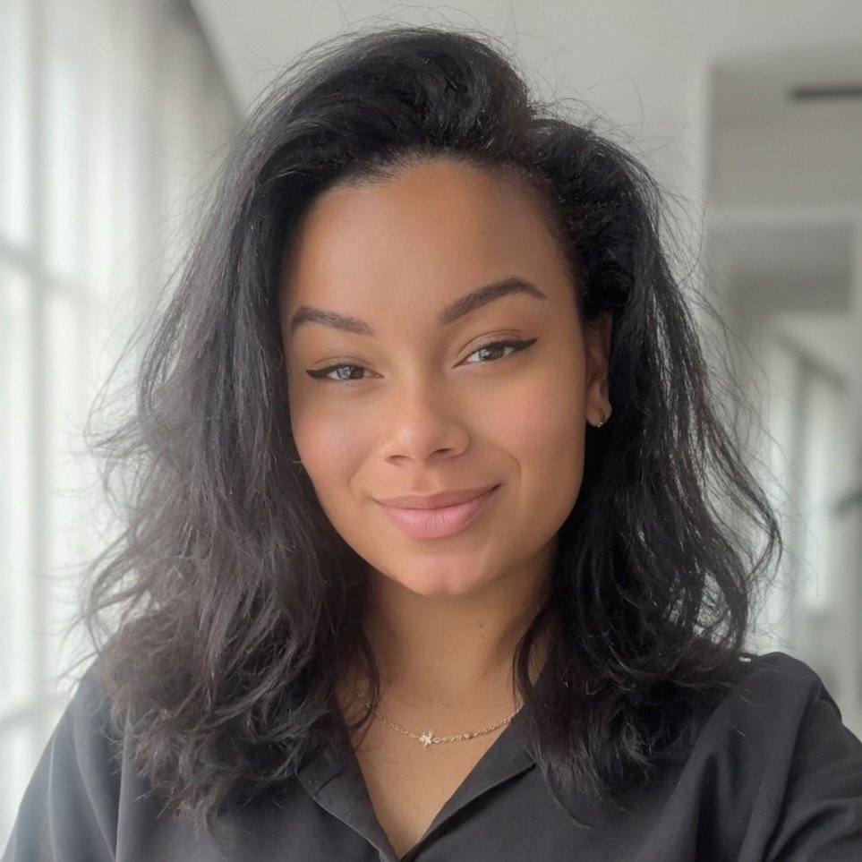A young woman with shoulder-length curly black hair, wearing a black shirt and a delicate gold necklace with a star pendant, smiling softly in a well-lit indoor space.