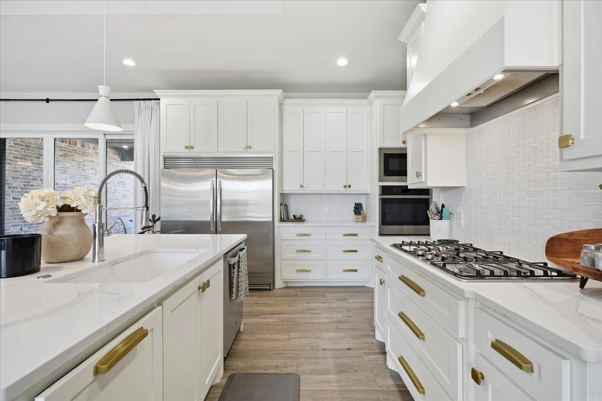 Modern white kitchen with gold hardware, stainless steel refrigerator, and a gas stove. Decor includes a vase with white flowers and a wooden tray.