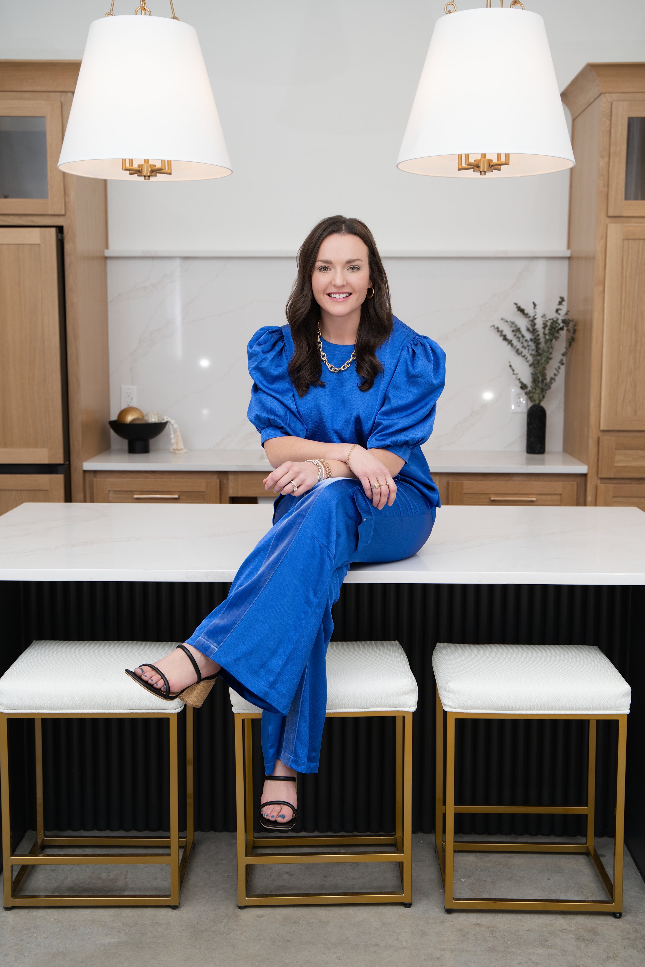 A woman with dark hair sitting on a white and gold stool in a modern kitchen with light wood cabinets, white marble backsplash, and two large white hanging pendant lights, wearing a bright blue outfit and black strappy sandals.