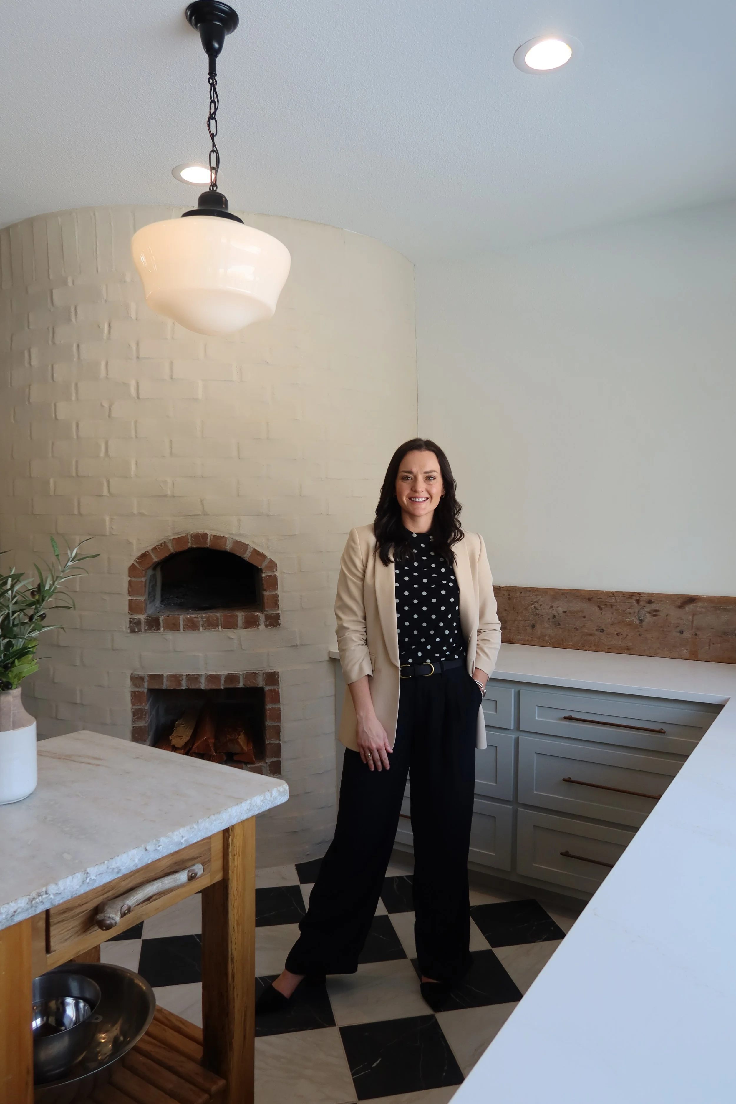 A woman with long dark hair smiling and standing in a kitchen. She is wearing a beige blazer over a black polka-dot blouse and black pants. The kitchen features a white brick fireplace, a chandelier, a potted plant, and gray cabinets with wooden handles.