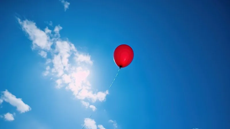 A red balloon floating in a blue sky with a few white clouds.