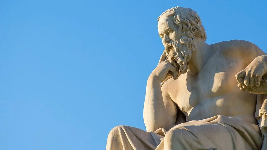 Close-up of the statue of a seated philosopher with one hand on his chin, against a blue sky background.