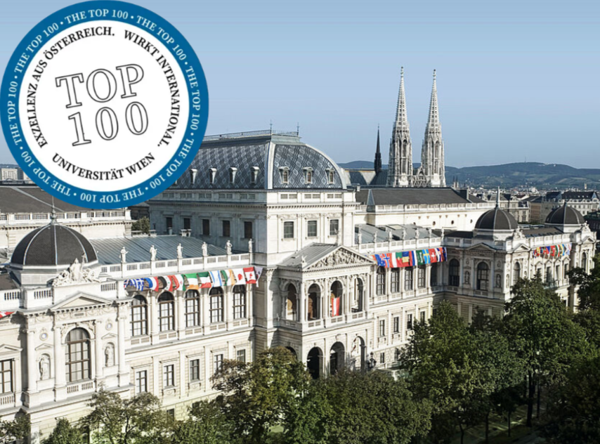 Aerial view of a grand historic building with flags hanging from its facade, two tall church spires in the background, and a city skyline with hills in the distance.
