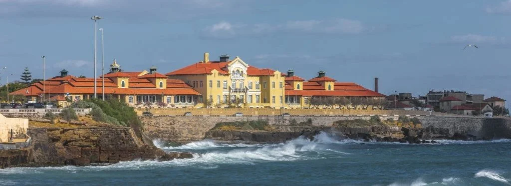 A historic yellow building with red roofs on a rocky coastline with waves crashing below, under a partly cloudy sky.